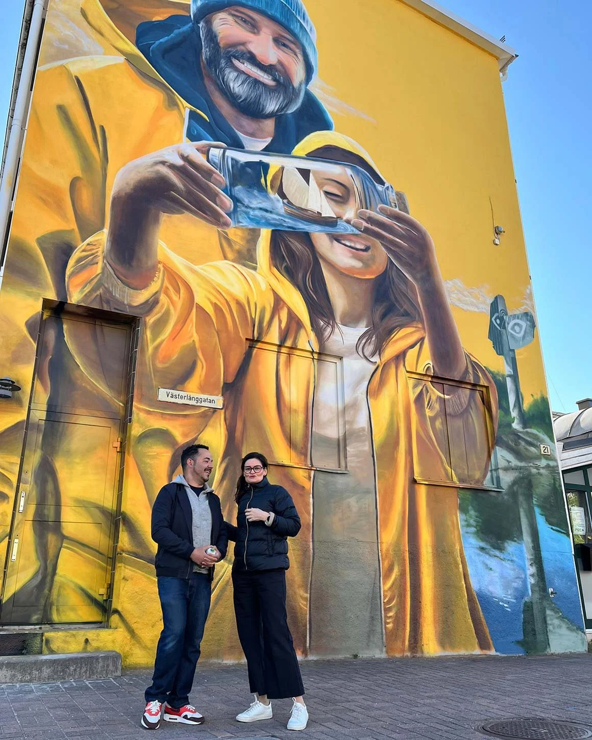 Två människor står framför en stor väggmålning av en man och en kvinna, där mannen håller i ett glas med en modell av en segelbåt och kvinnan guidar glaset till munnen, båda bär gula jackor.