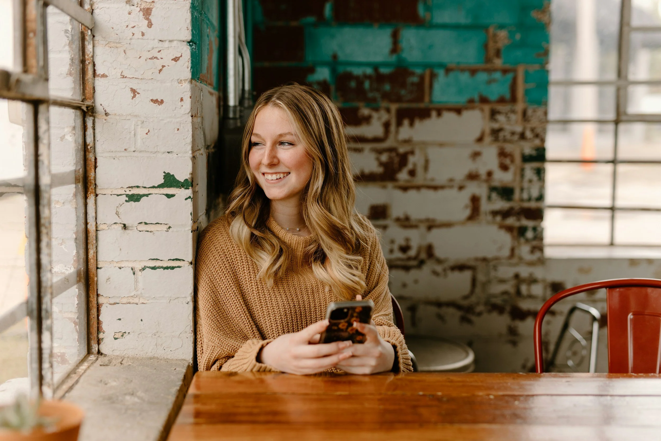 A young woman with wavy blonde hair, wearing a tan sweater, smiling and holding a smartphone, sitting at a wooden table in a rustic, industrial-style cafe with exposed brick walls and large windows.