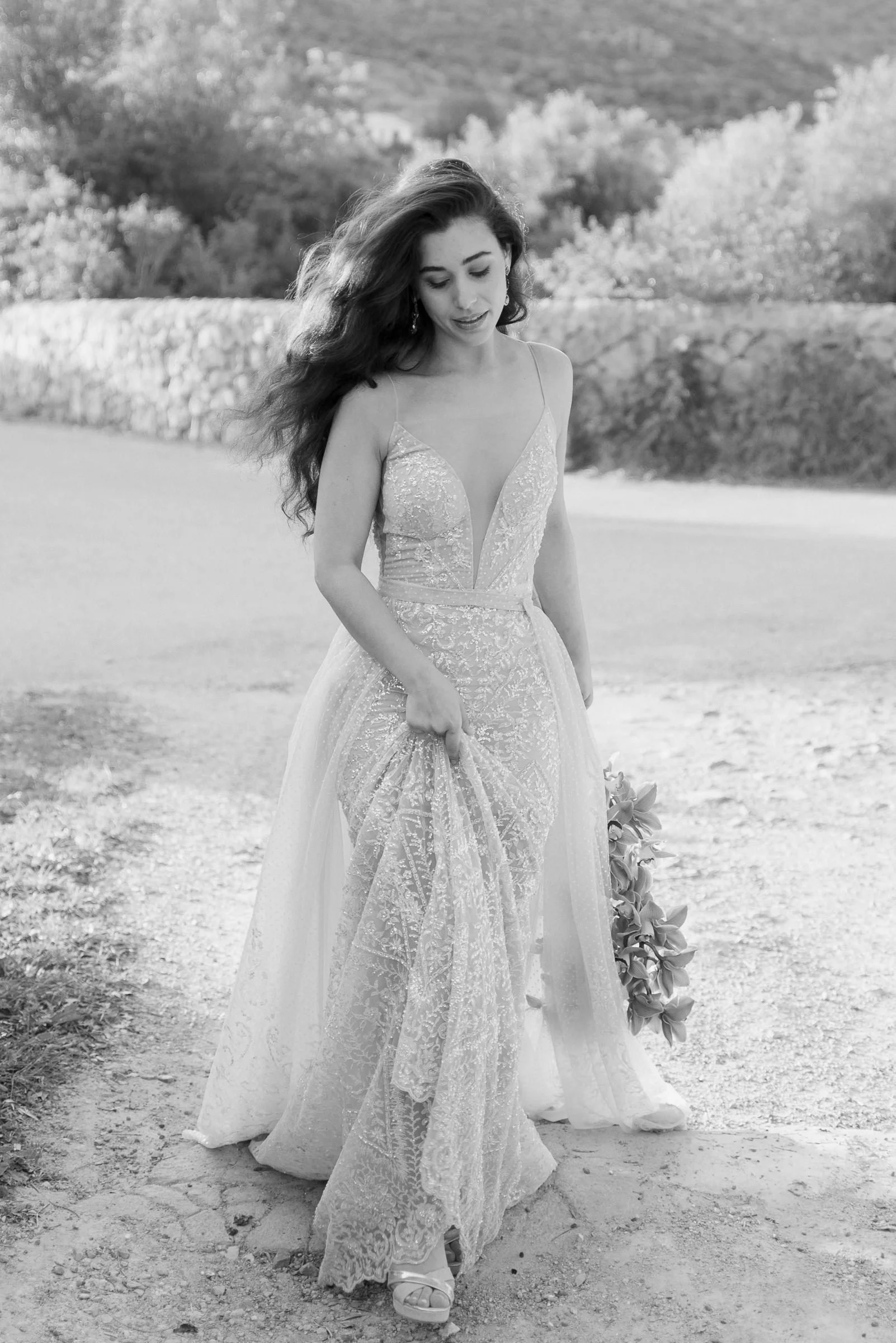A woman in a wedding dress walking outdoors on a dirt path with a stone wall and trees in the background.