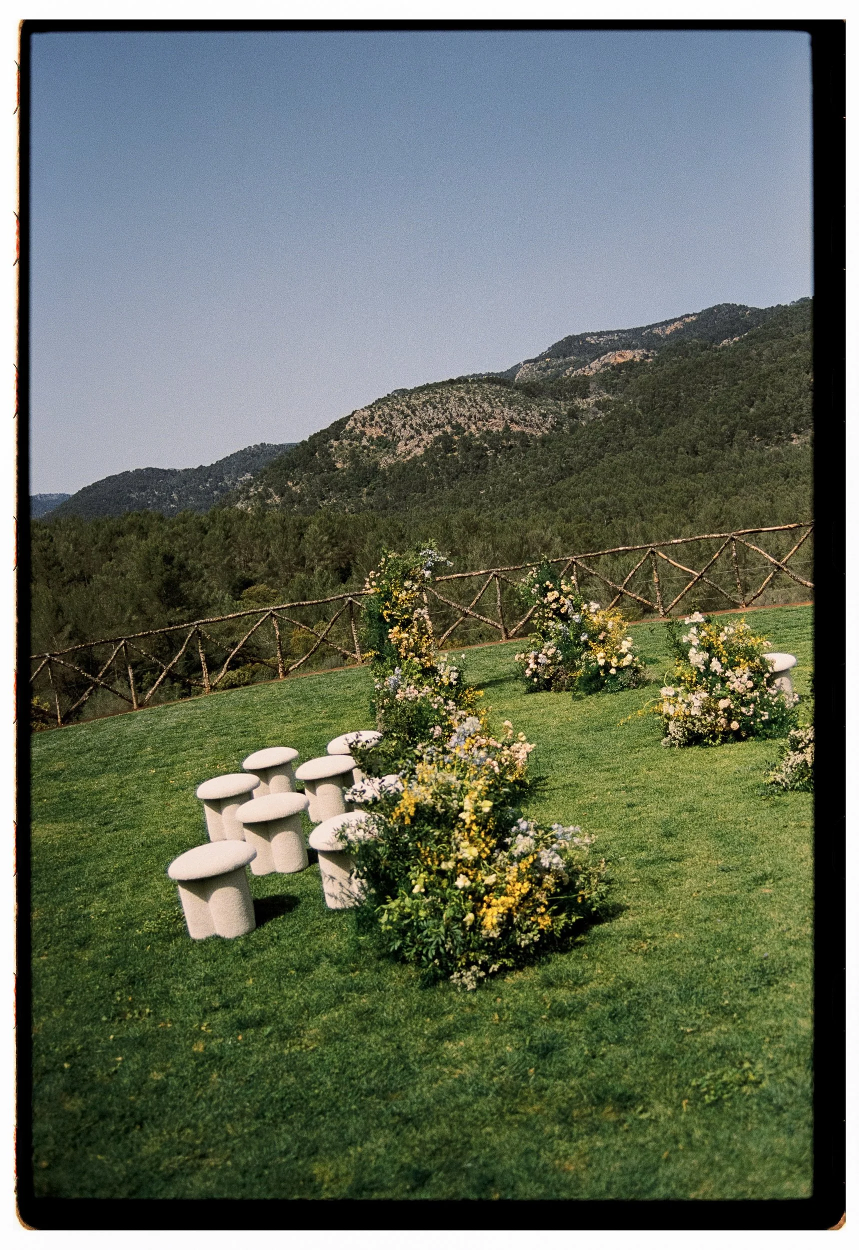 Outdoor scene with a grassy lawn, white mushroom-shaped chairs, flower arrangements, wooden fence, forested mountains in the background, and clear sky.