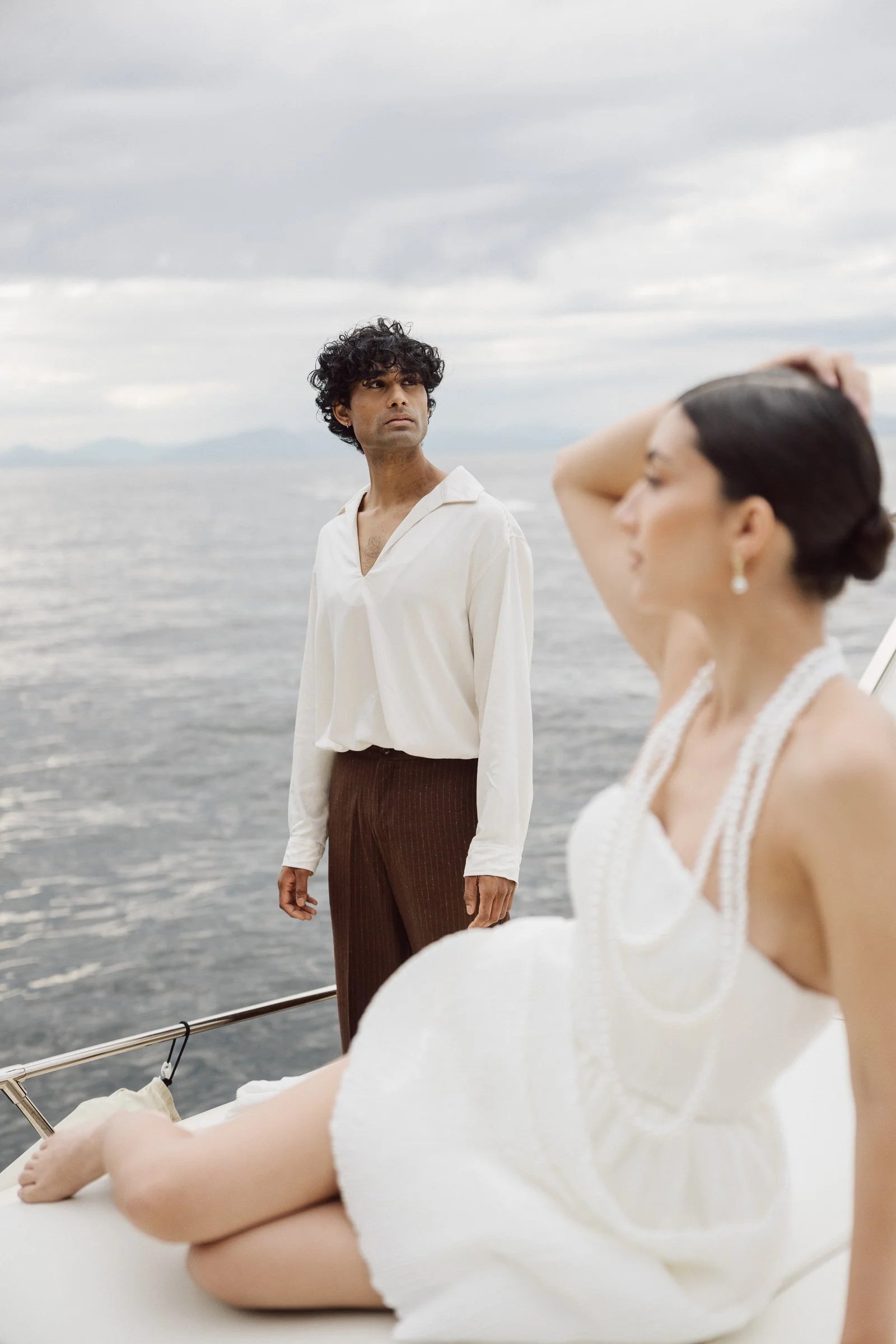 A man with dark curly hair and a serious expression standing on a boat near the ocean, while a woman with dark hair in a bun, wearing a white dress and pearl earrings, is in the foreground with her arm raised over her head against a cloudy sky.