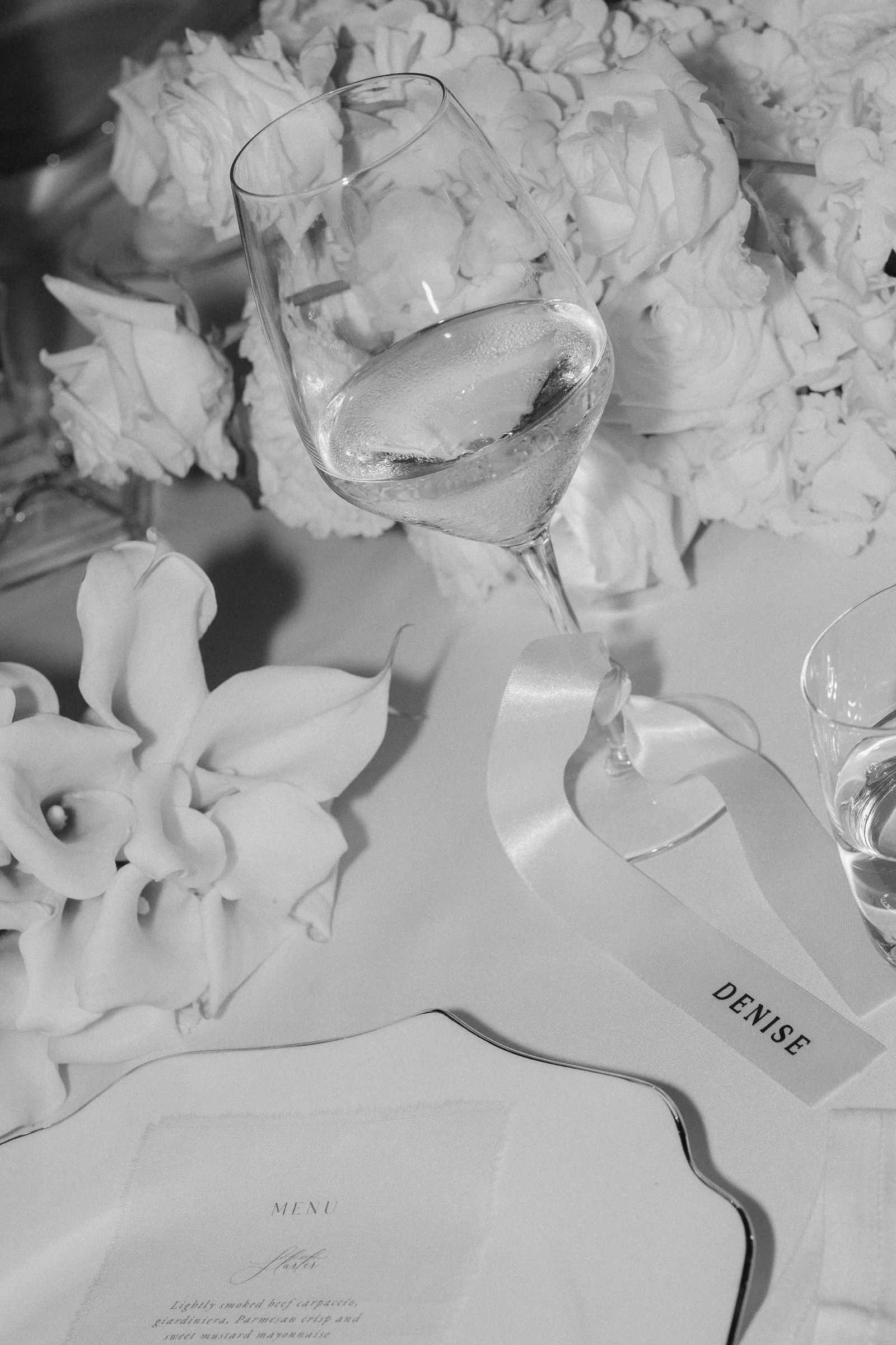 A glass of sparkling water or champagne with ice cubes placed at an elegant event table, surrounded by flower decorations and a menu card with the name 'Denise' visible.