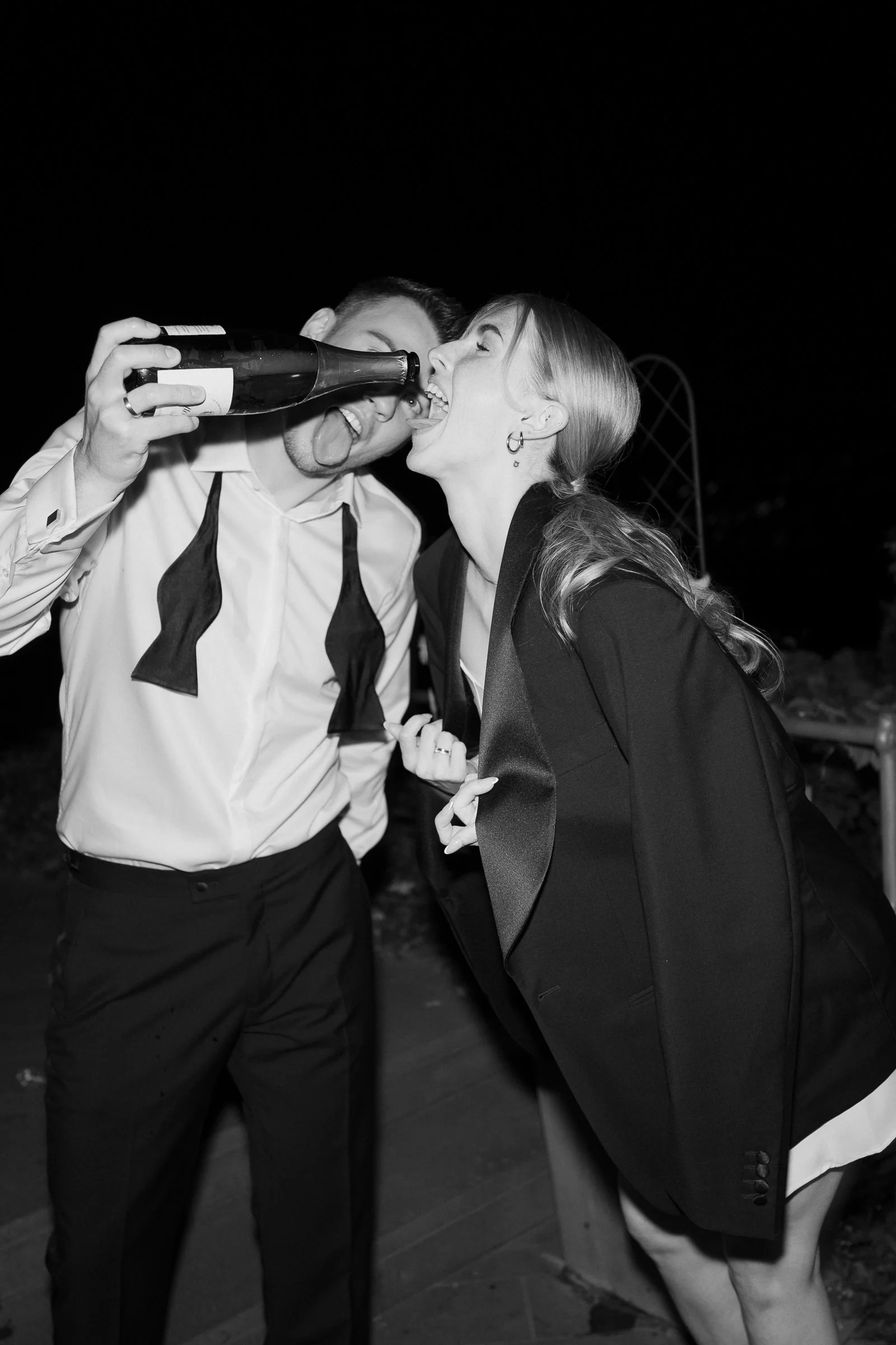 Two people, a man and a woman, are having fun at a party. The man is holding a bottle and pretending to feed the woman with it. Both are smiling and appear to be enjoying themselves. The background is dark, indicating nighttime or an indoor setting.