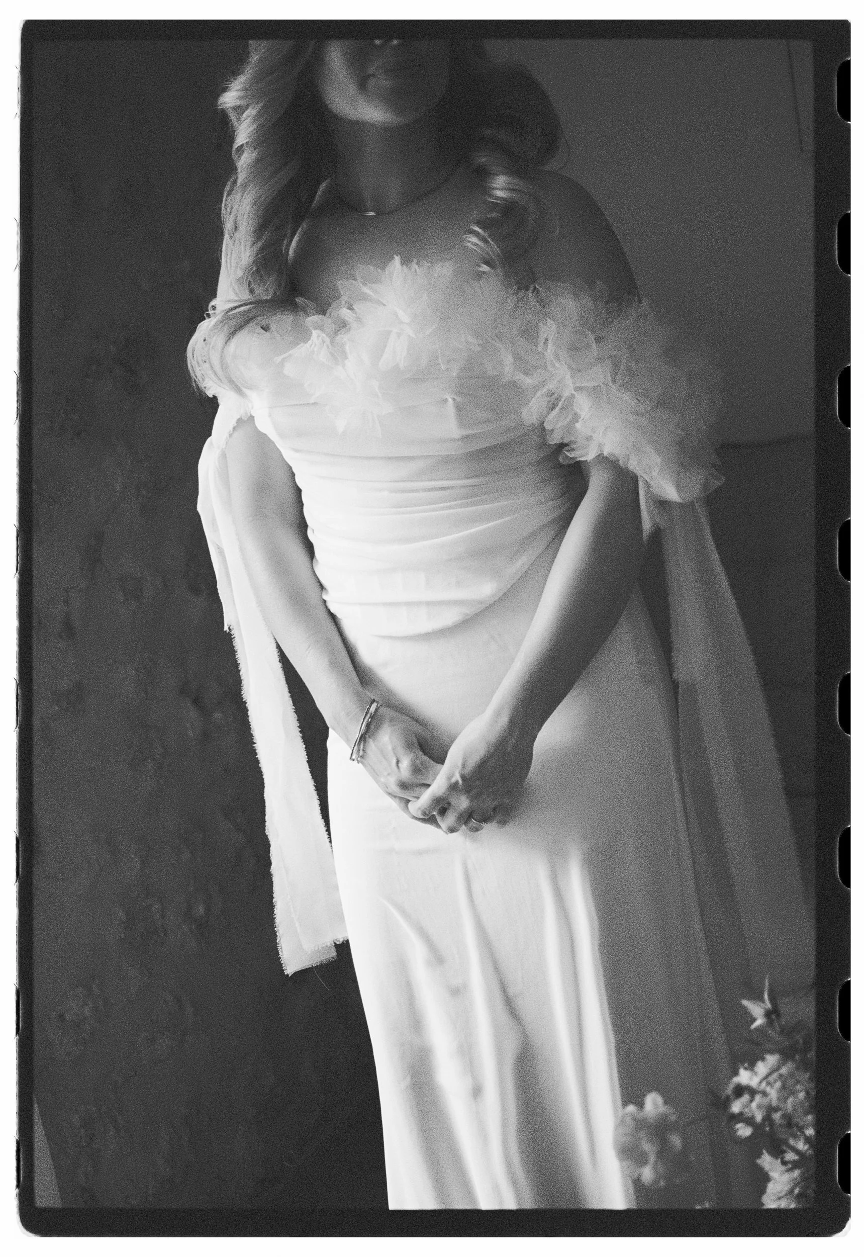 Black and white photograph of a woman in a wedding dress, holding a bouquet, standing outdoors with trees in the background.