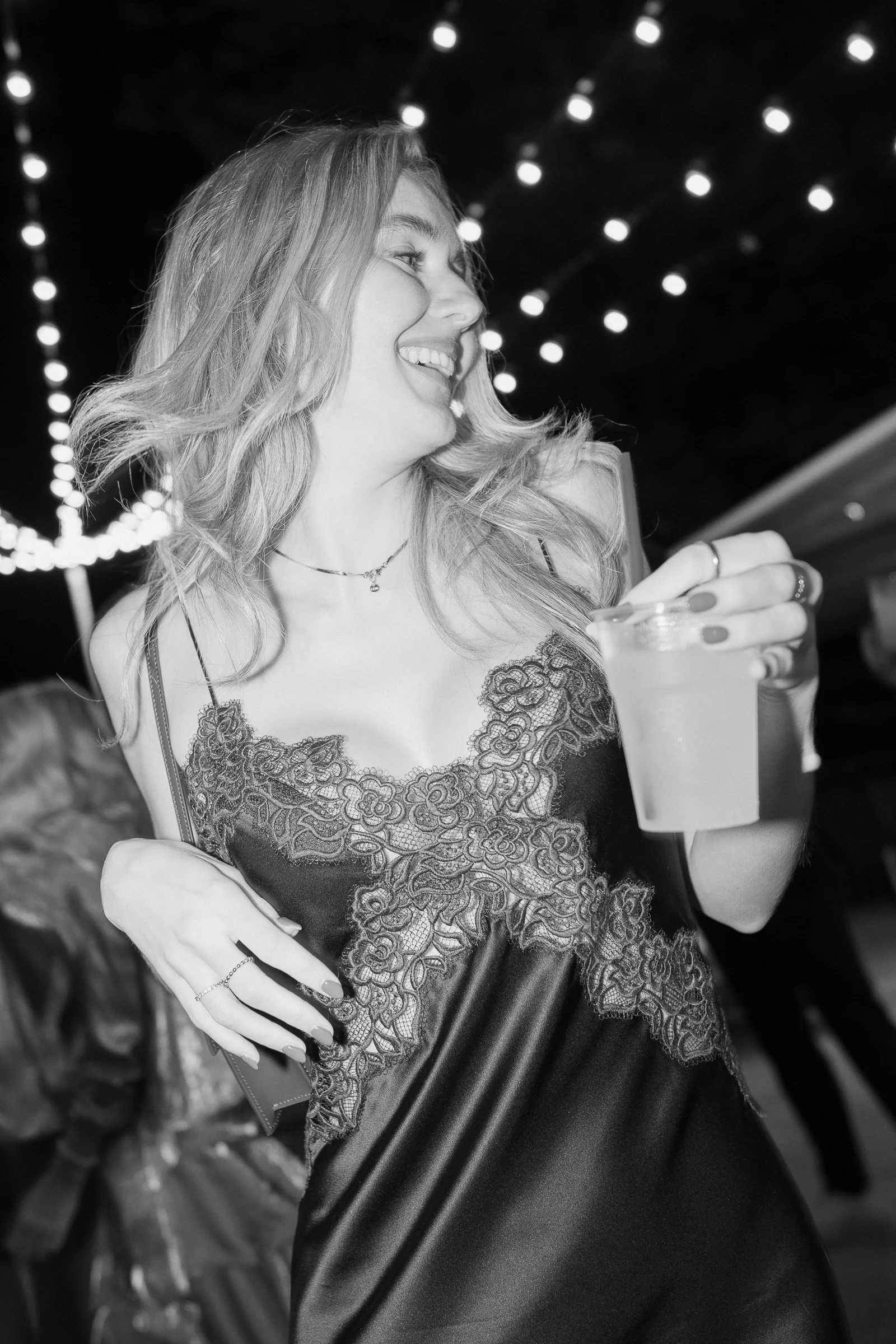 A woman smiling, holding a drink, wearing a dark lace dress, with long wavy hair, in a setting with string lights and blurred background.