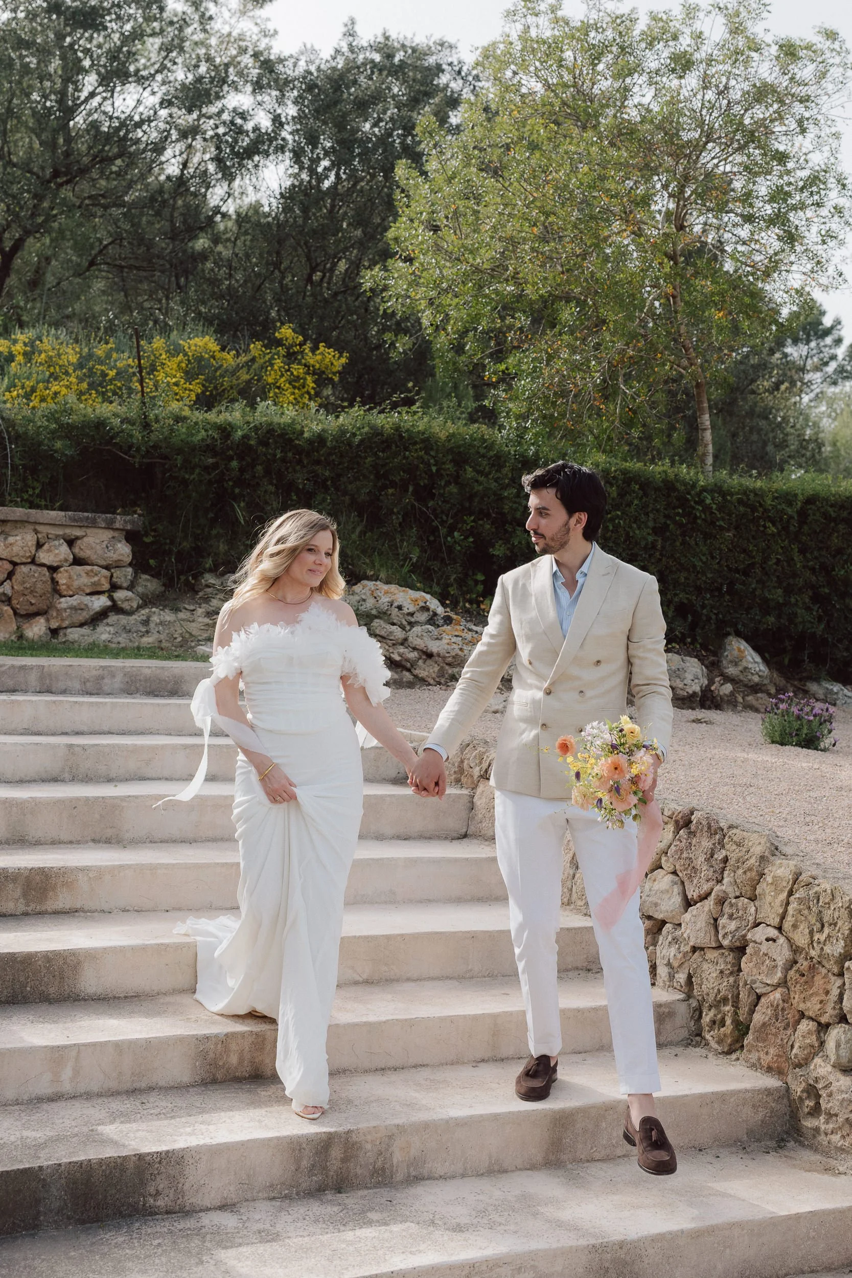 A couple holding hands and walking down outdoor stone steps, with a woman in a white wedding dress and a man in a cream-colored suit, holding a bouquet of flowers, surrounded by greenery and trees.