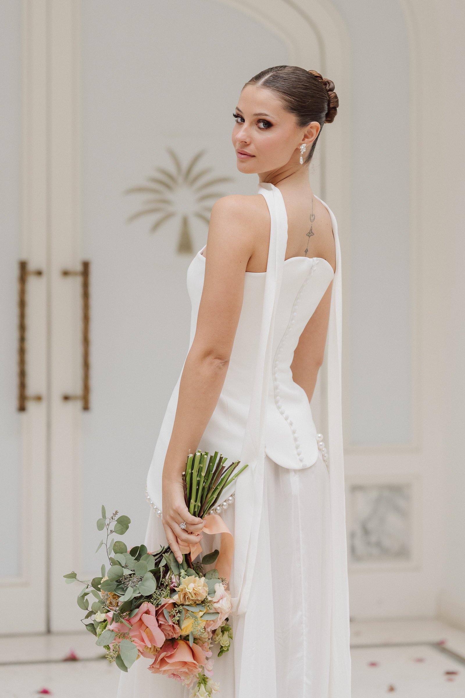 A woman in a white wedding dress holding a bouquet of flowers, with her hair styled in an elegant updo, standing indoors with a subtle background.