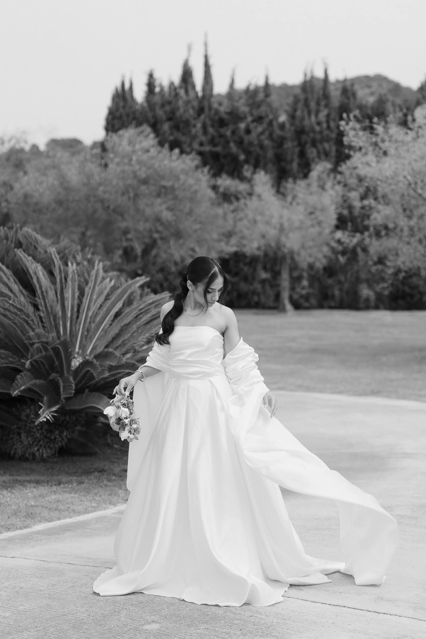 A woman in a white wedding gown holding a bouquet in an outdoor setting with trees and bushes in the background.
