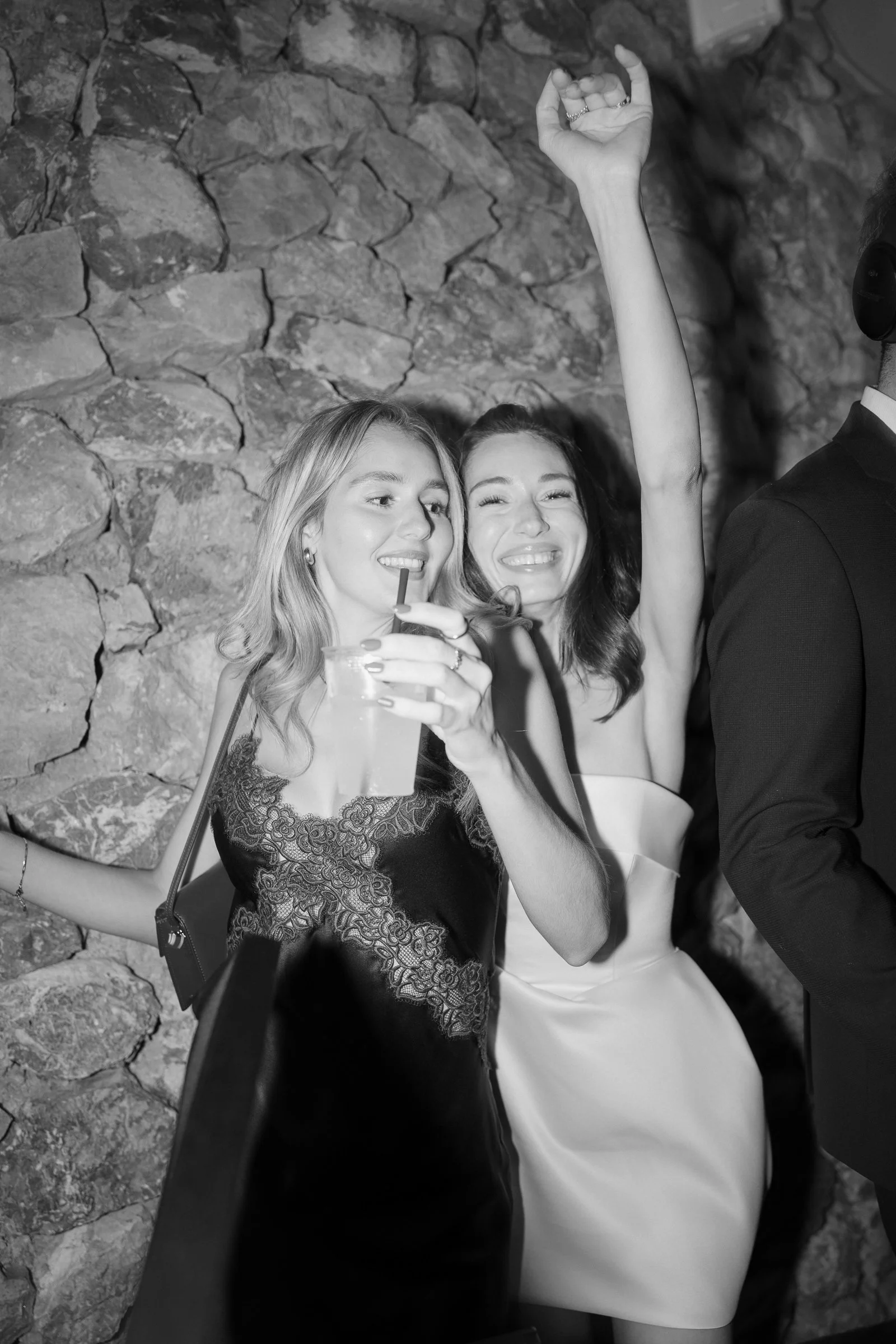 Two women in elegant dresses dancing and smiling, with one making a hand gesture and the other holding a drink, against a stone wall background.