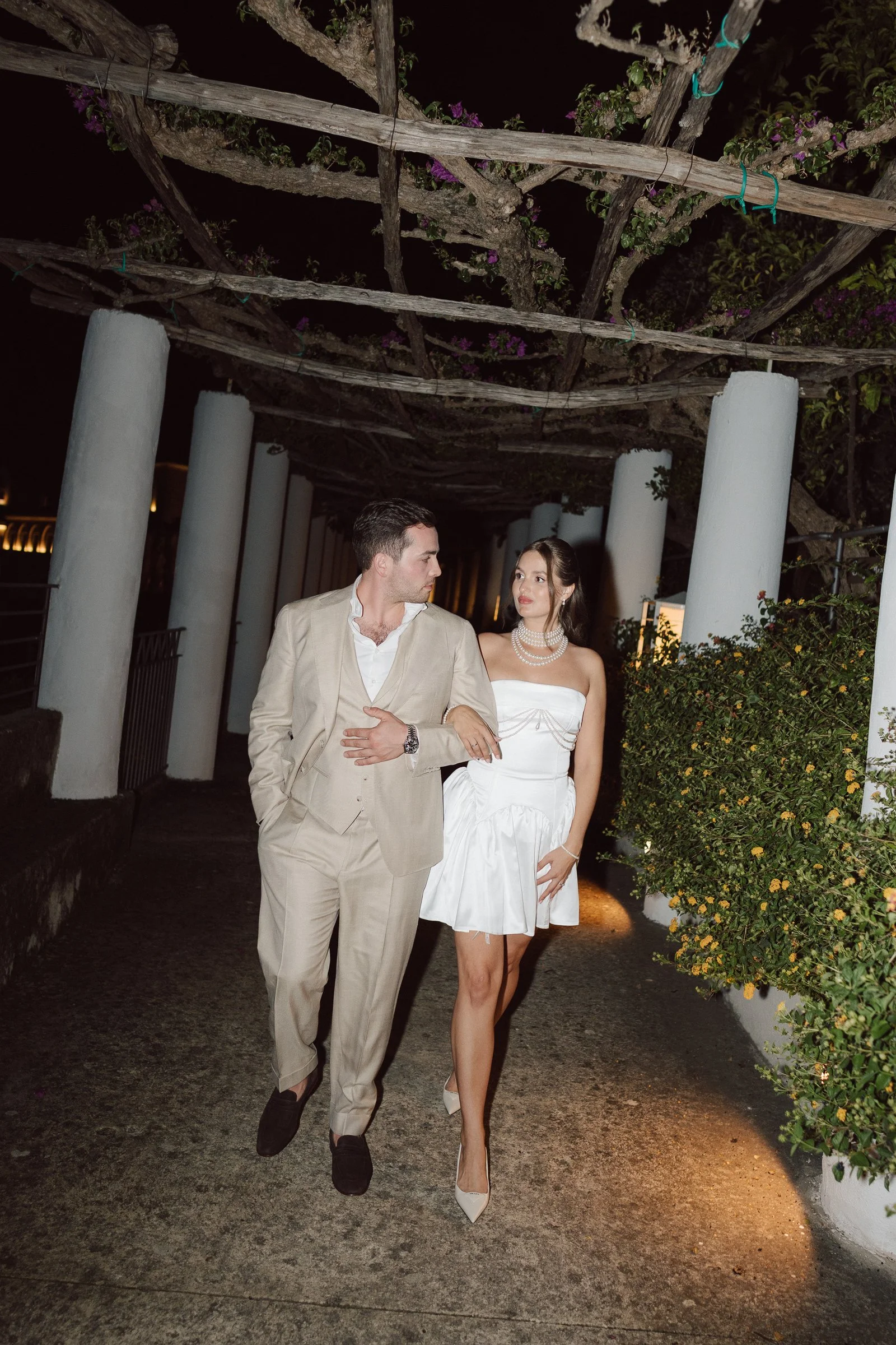 A man and woman dressed in formal attire walking outdoors at night, surrounded by tall white columns and greenery, under a wooden overhead structure with purple flowers.