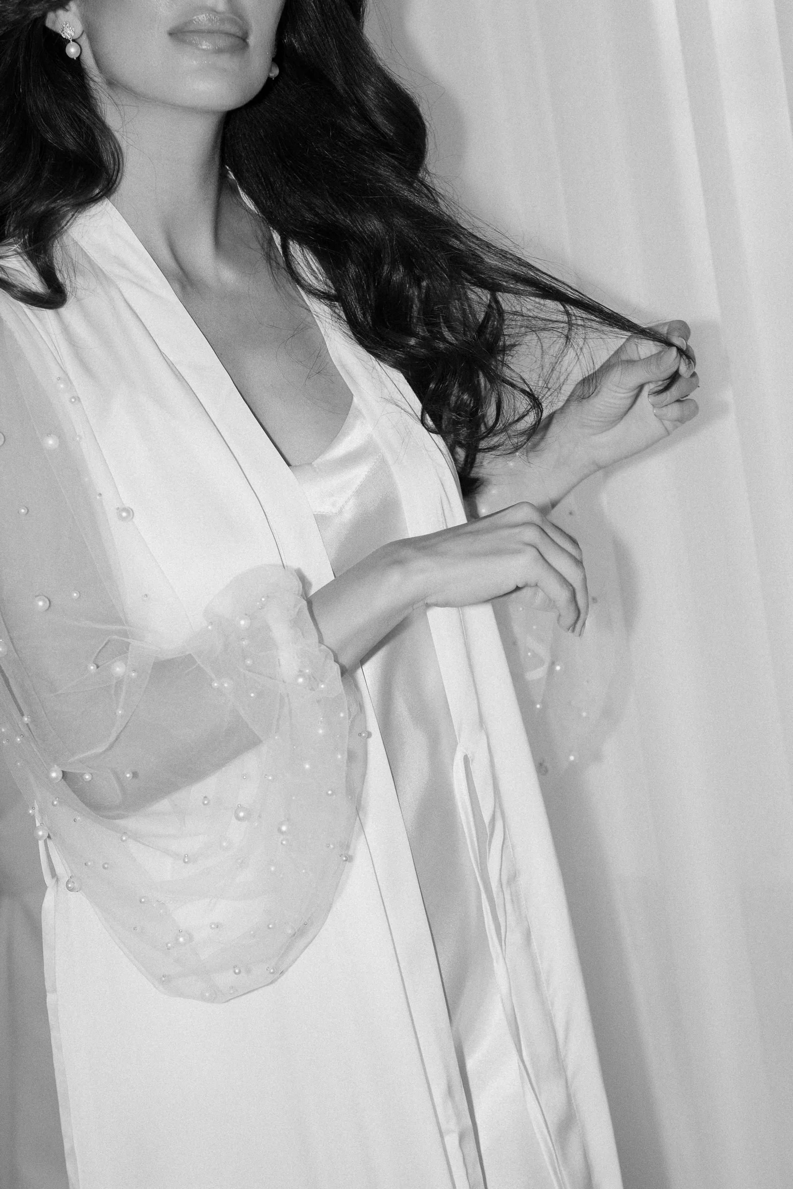 A black-and-white photo of a woman with long wavy hair wearing a white satin dress and pearl earrings, sitting at a table with her hands clasped.