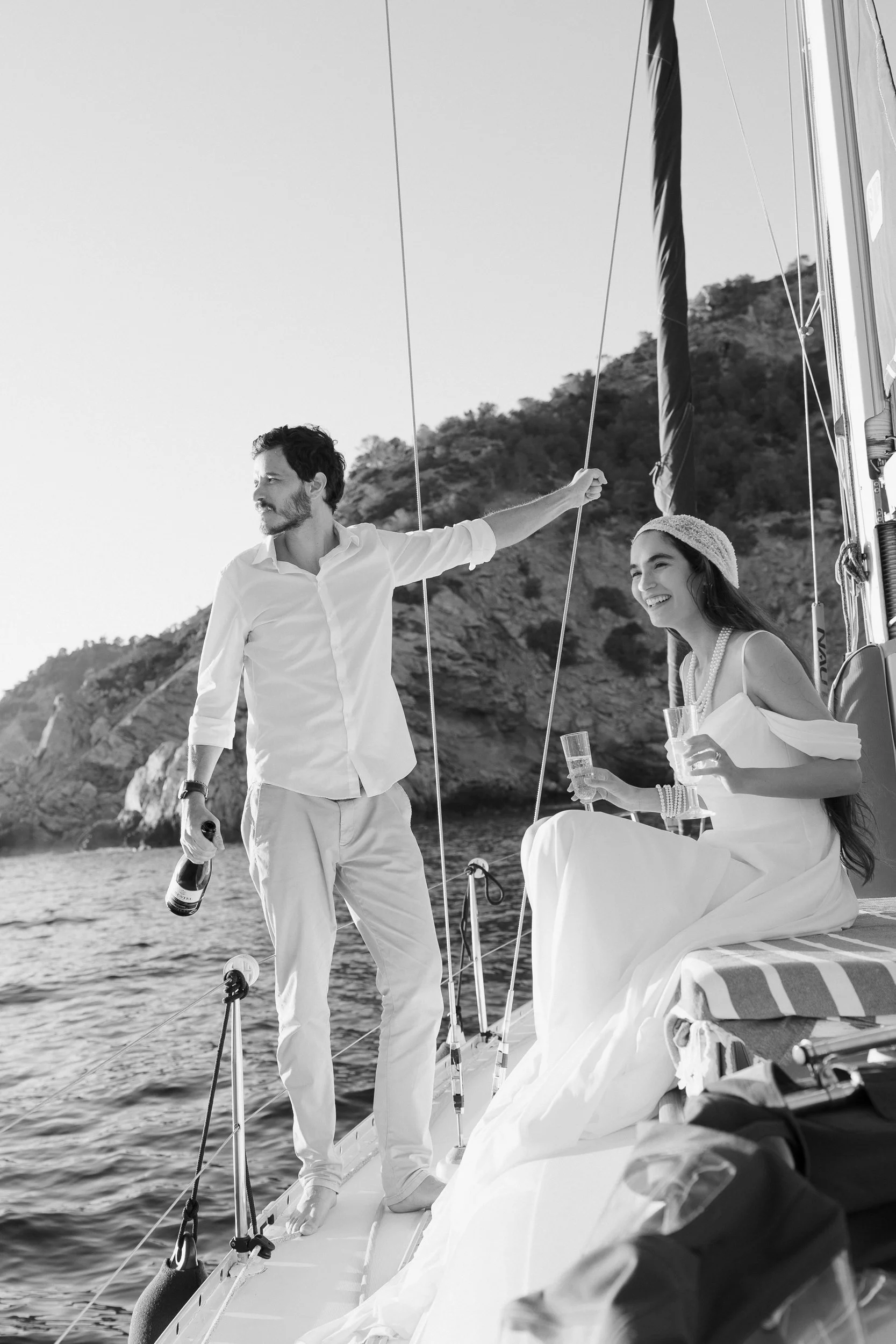 A man and woman on a sailboat enjoying a sunny day by the water, with the woman sitting and the man standing holding a bottle, both dressed in light-colored clothing, near a scenic hillside.