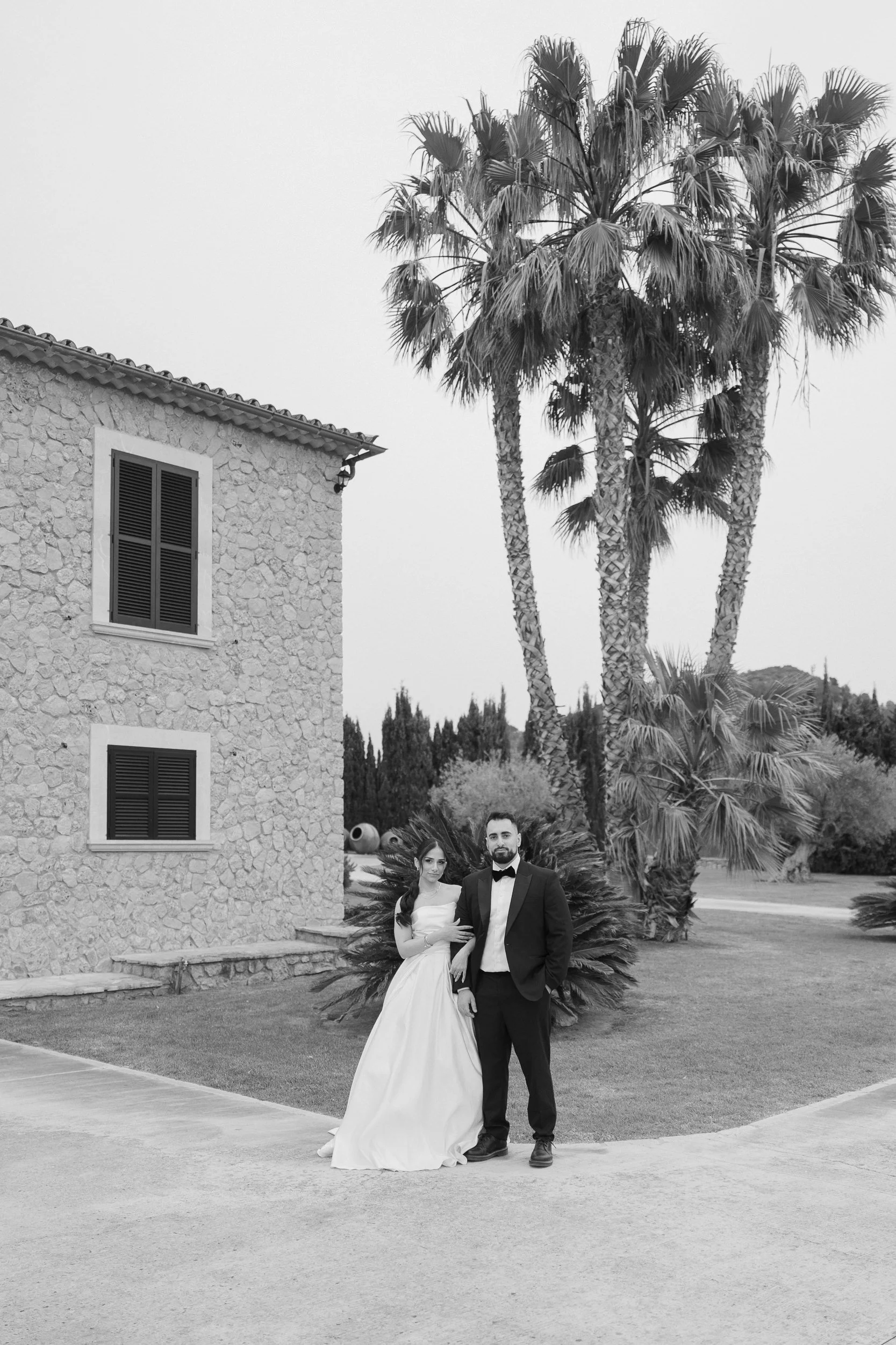 A black and white photo of a couple dressed in wedding attire, standing outdoors in front of a stone building with palm trees in the background.