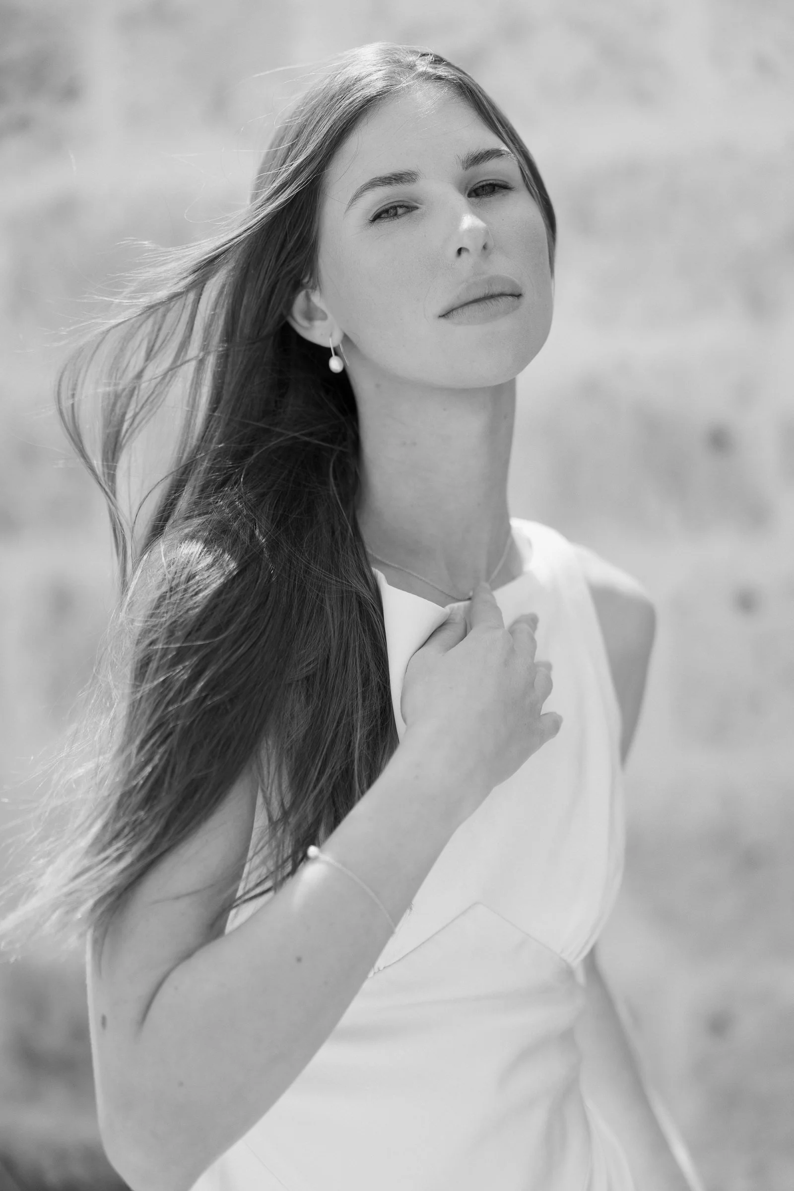 Close-up black and white portrait of a young woman with long flowing hair, wearing a pearl earring, looking directly at the camera with a confident expression. Background is blurred.