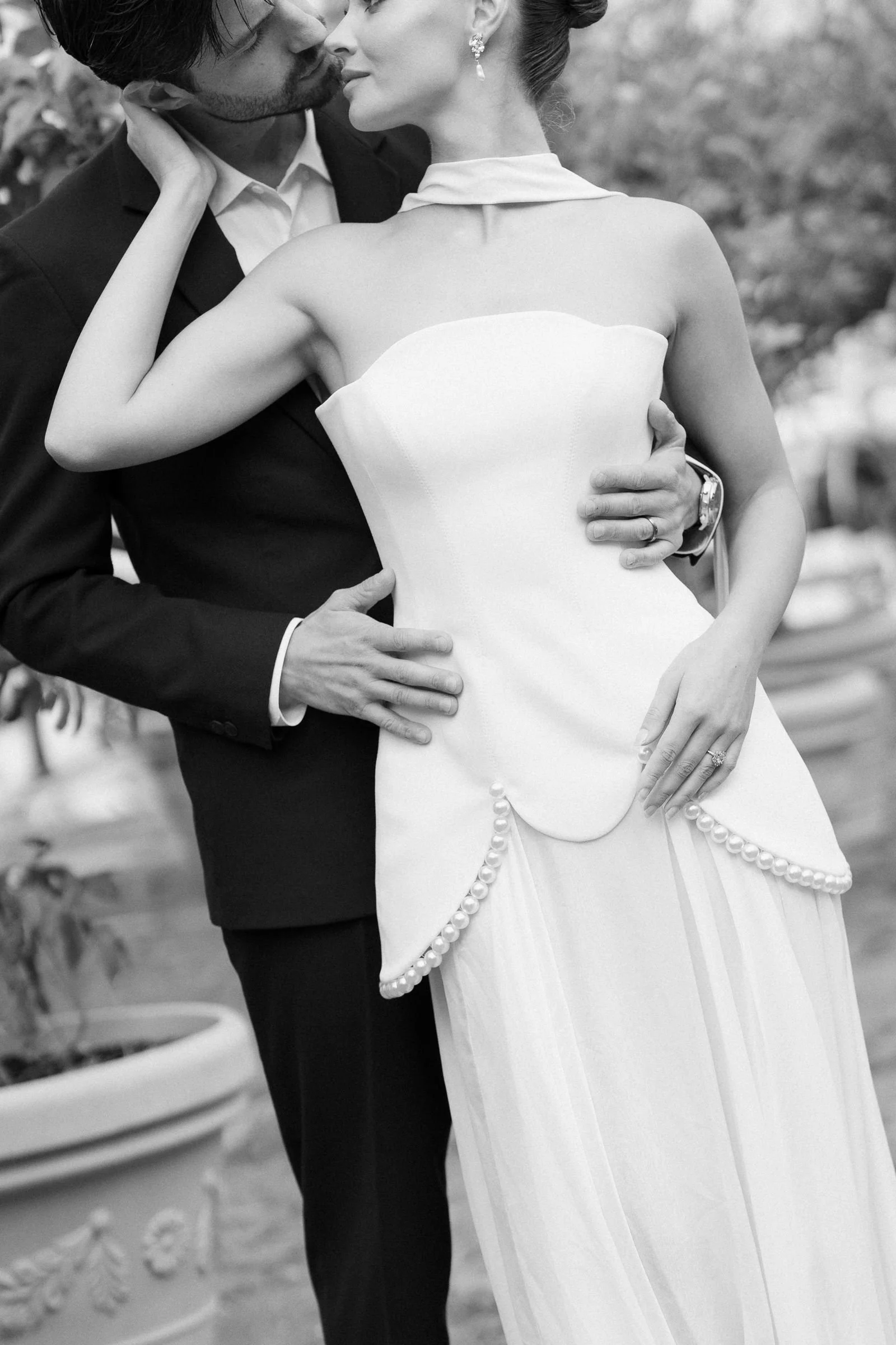A black and white photo of a couple kissing, the woman wearing a strapless dress with pearl embellishments and earrings, the man in a tuxedo, holding each other closely.
