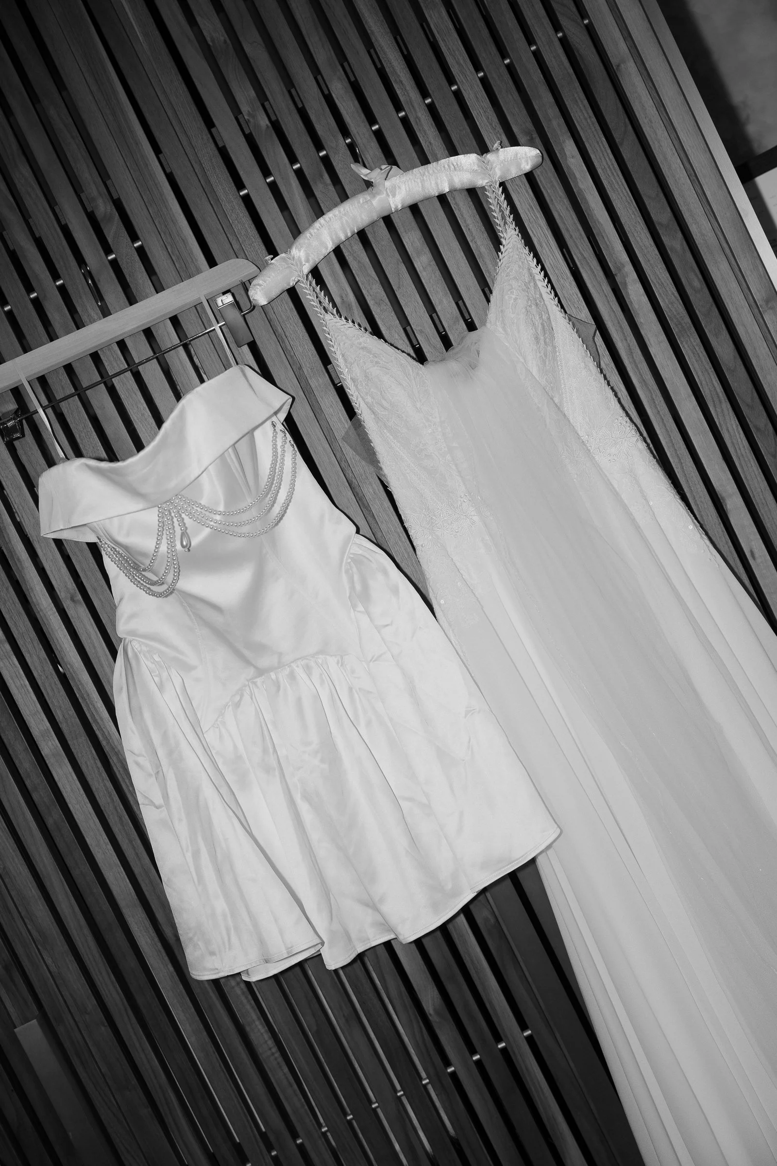 A wedding dress hanging on a padded hanger and a satin dress with pearl embellishments hanging on a wooden rack against a wooden slat wall.