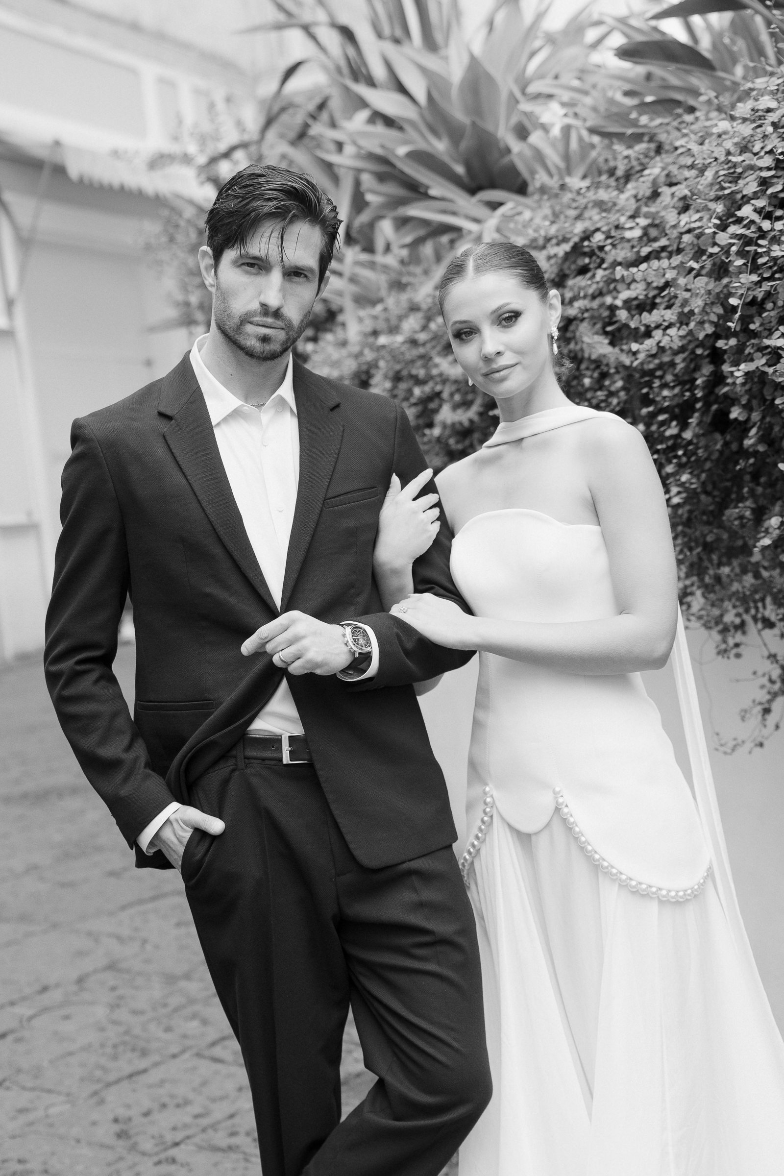 Black and white photo of a man in a suit and a woman in a wedding dress standing outdoors, with lush foliage in the background.