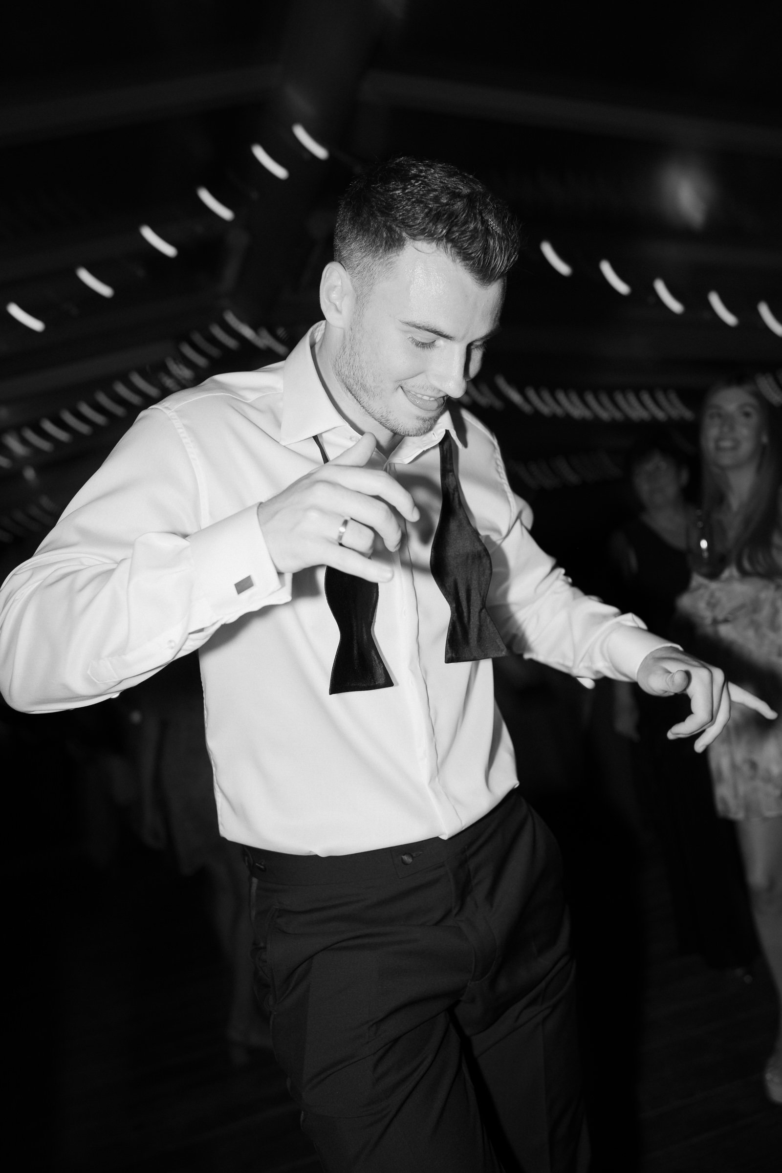 A young man dressed in a white shirt and black pants, holding a wine glass, smiling, at a social event or party, with a woman in the background smiling and looking towards him.