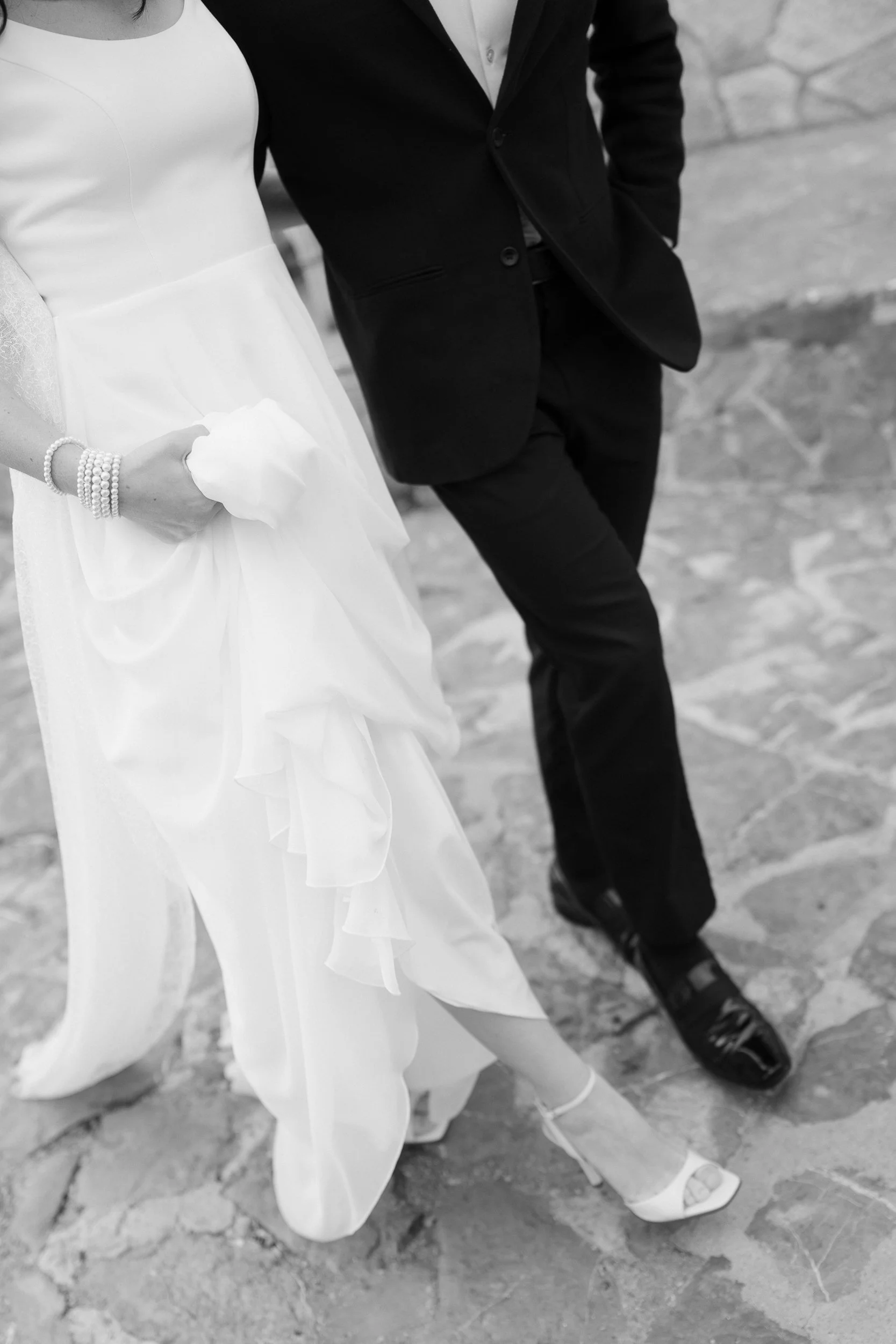 Black and white photo of a bride and groom standing on a stone pathway. The bride is wearing a white gown and holding a tissue, and the groom is in a black suit with his hands in his pockets. Both are wearing formal shoes.
