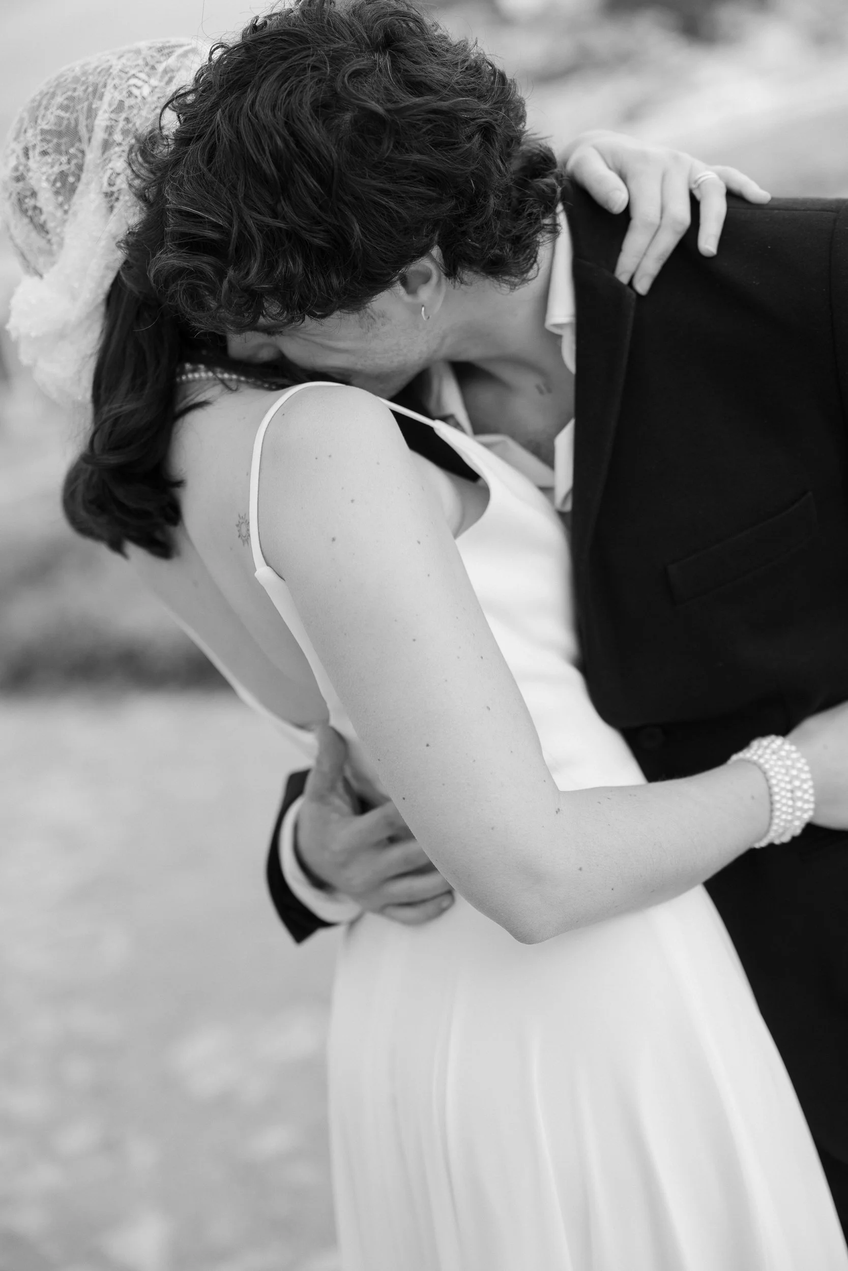 A black and white photograph of a couple sharing a kiss at their wedding, with the bride in a sleeveless dress and the groom in a suit, with a blurred outdoor background.
