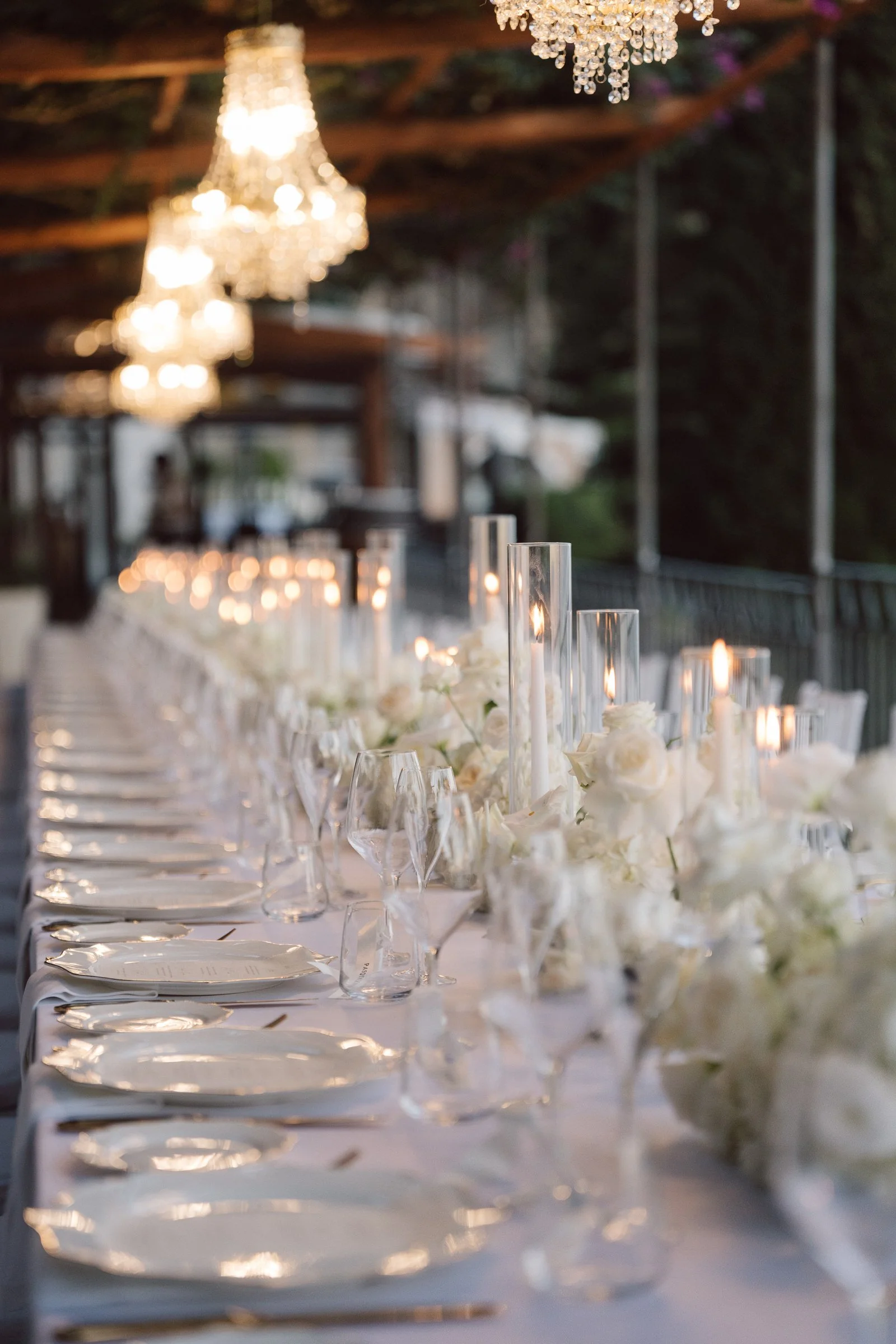 A banquet table decorated with white flowers, tall glass candle holders, and lit candles underneath a chandelier, likely set for an elegant event or wedding reception.