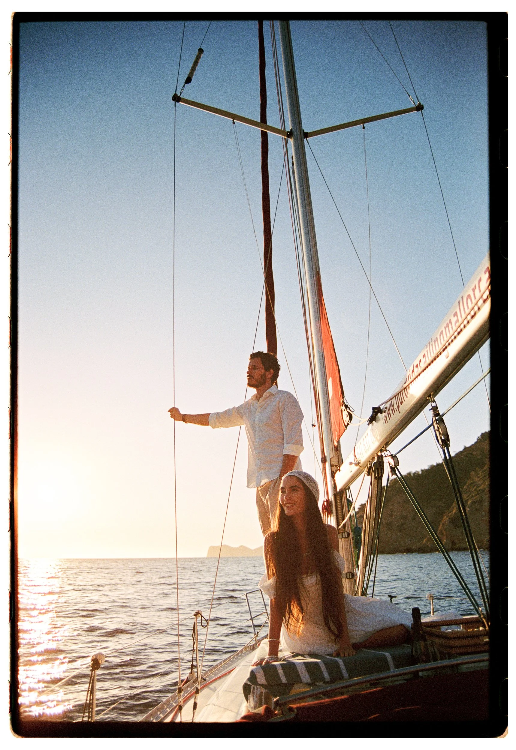 A man and a woman on a sailboat at sunset, with the man standing near the mast and the woman sitting on the deck, both smiling, with water and a distant island in the background.