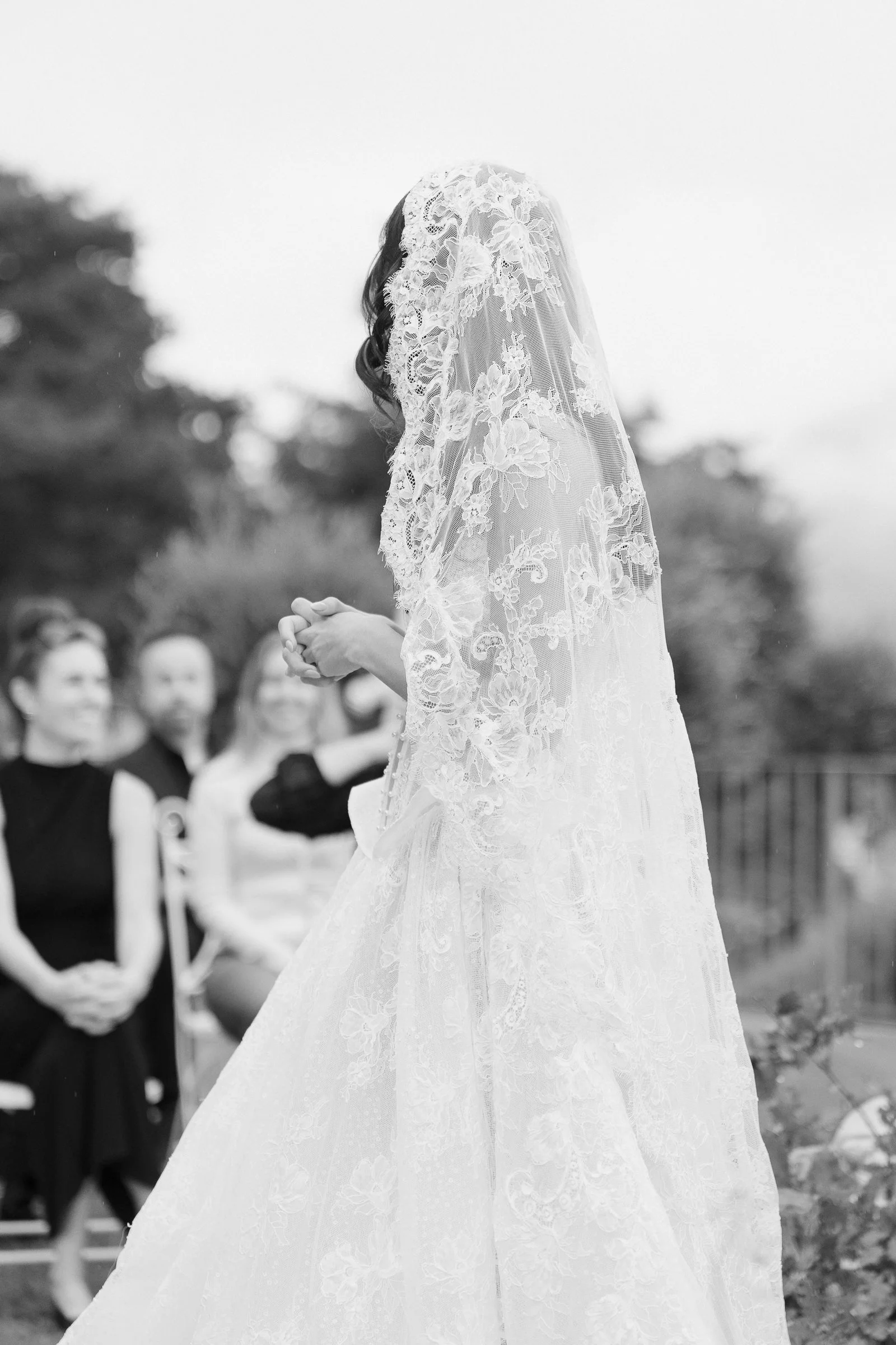 A bride in a lace wedding dress and veil is embracing or praying outdoors, with guests seated and watching in the background.