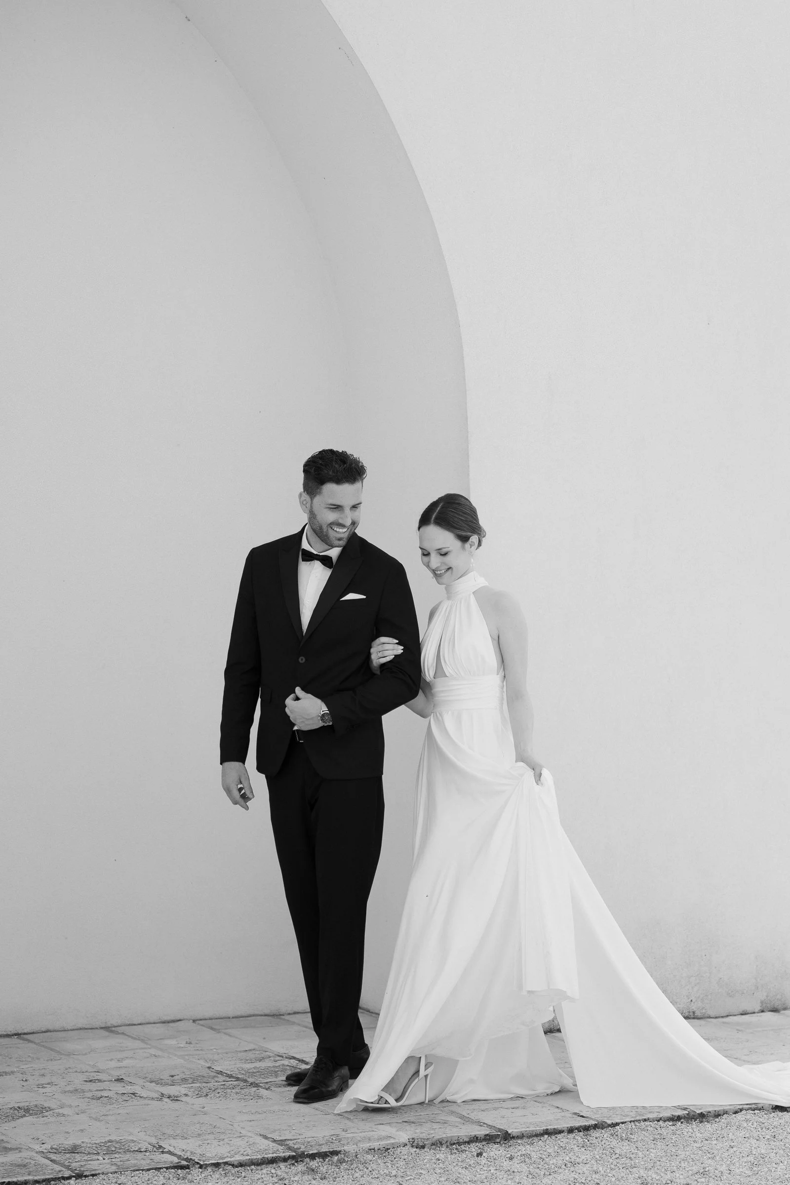 Black and white photo of a man and woman in wedding attire walking together on a sidewalk; the man is wearing a tuxedo, and the woman is in a white wedding dress.