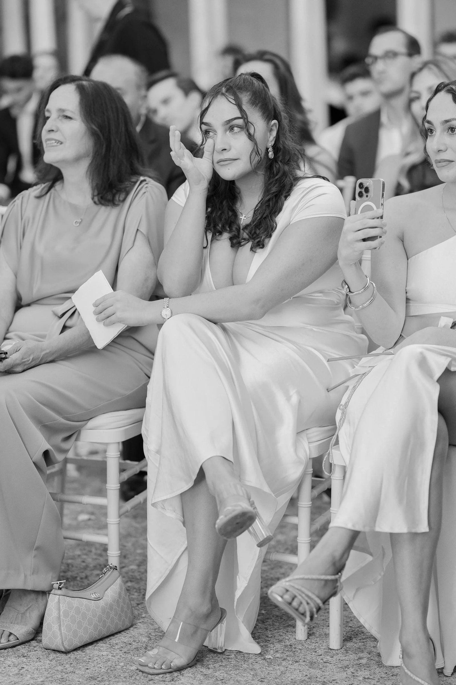 A group of women sitting and attending an event, with some holding phones, in formal attire, in black and white.