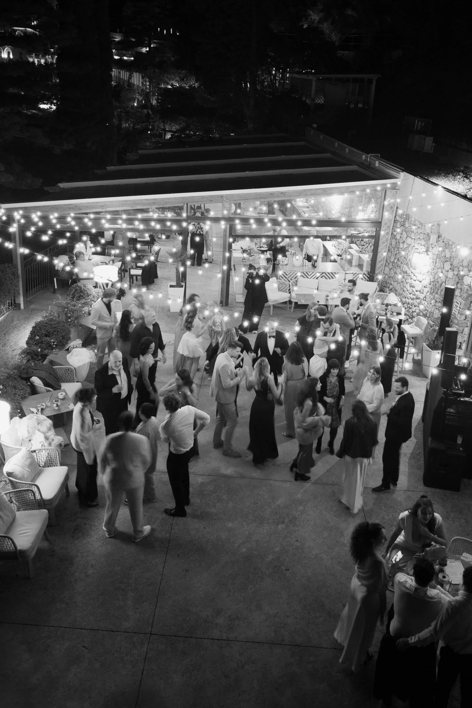 Nighttime outdoor celebration with people dancing and socializing under string lights, featuring an open patio with furniture and a lit building in the background.