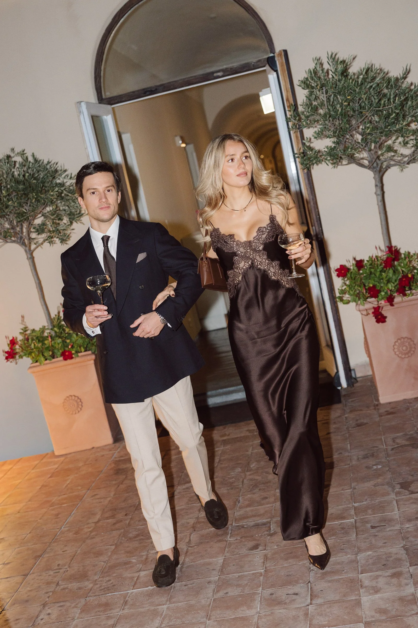 A man and a woman dressed in formal attire stand in front of an open doorway with potted plants on either side. The man wears a black suit with a white shirt and a dark tie, holding a glass of wine, while the woman wears a satin dark brown, satin sli