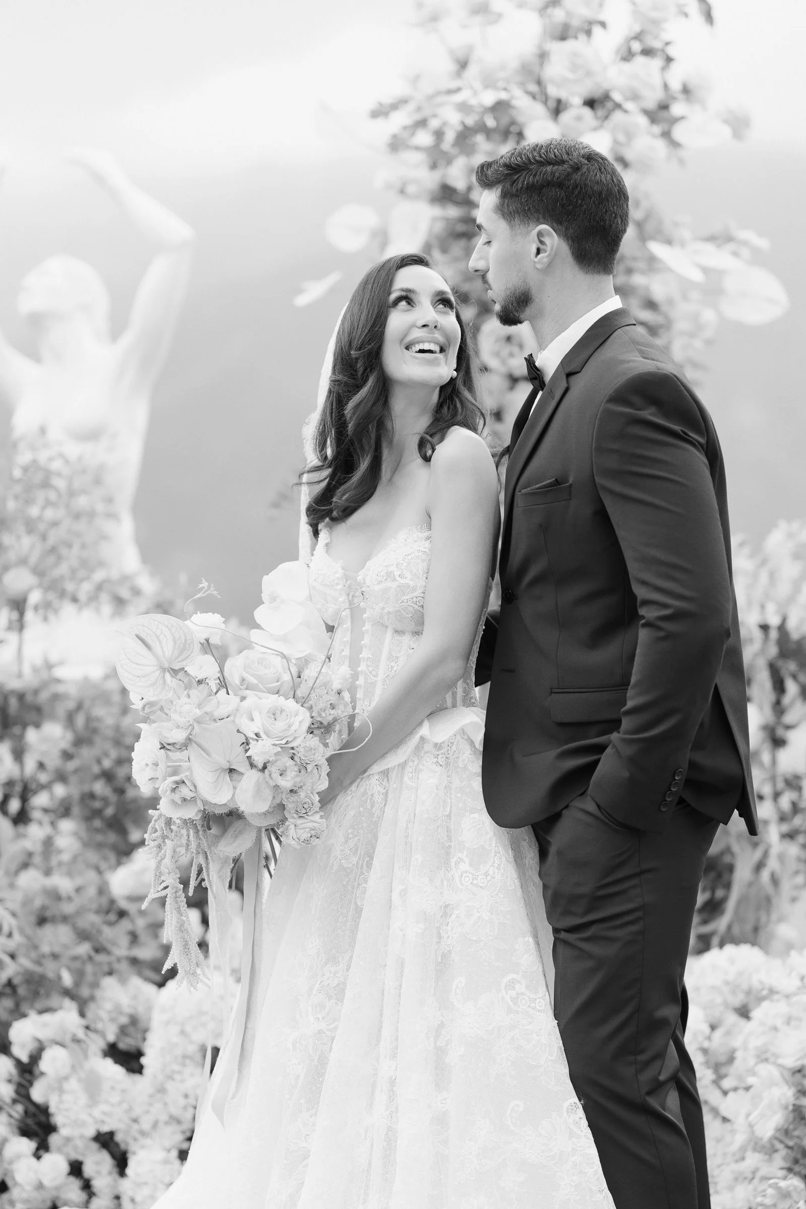 Black and white photo of a bride and groom during their wedding ceremony, standing close together with a background of flowers and a statue.