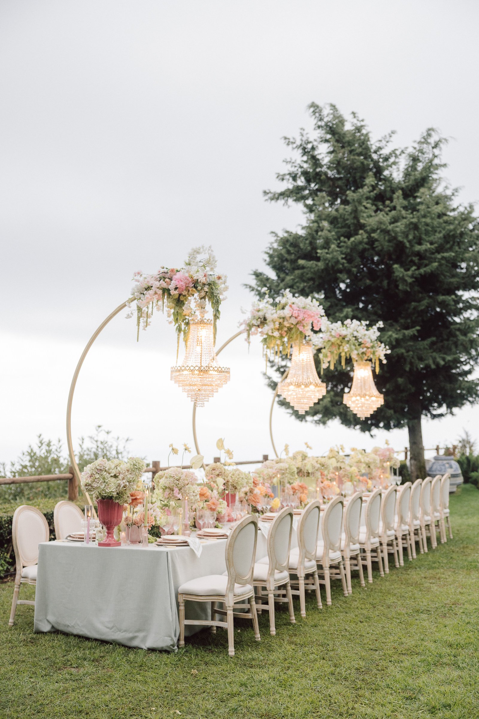 An outdoor wedding reception setup with a long banquet table decorated with floral arrangements, surrounded by white chairs, and hanging lantern-style lights with floral accents under a large tree.