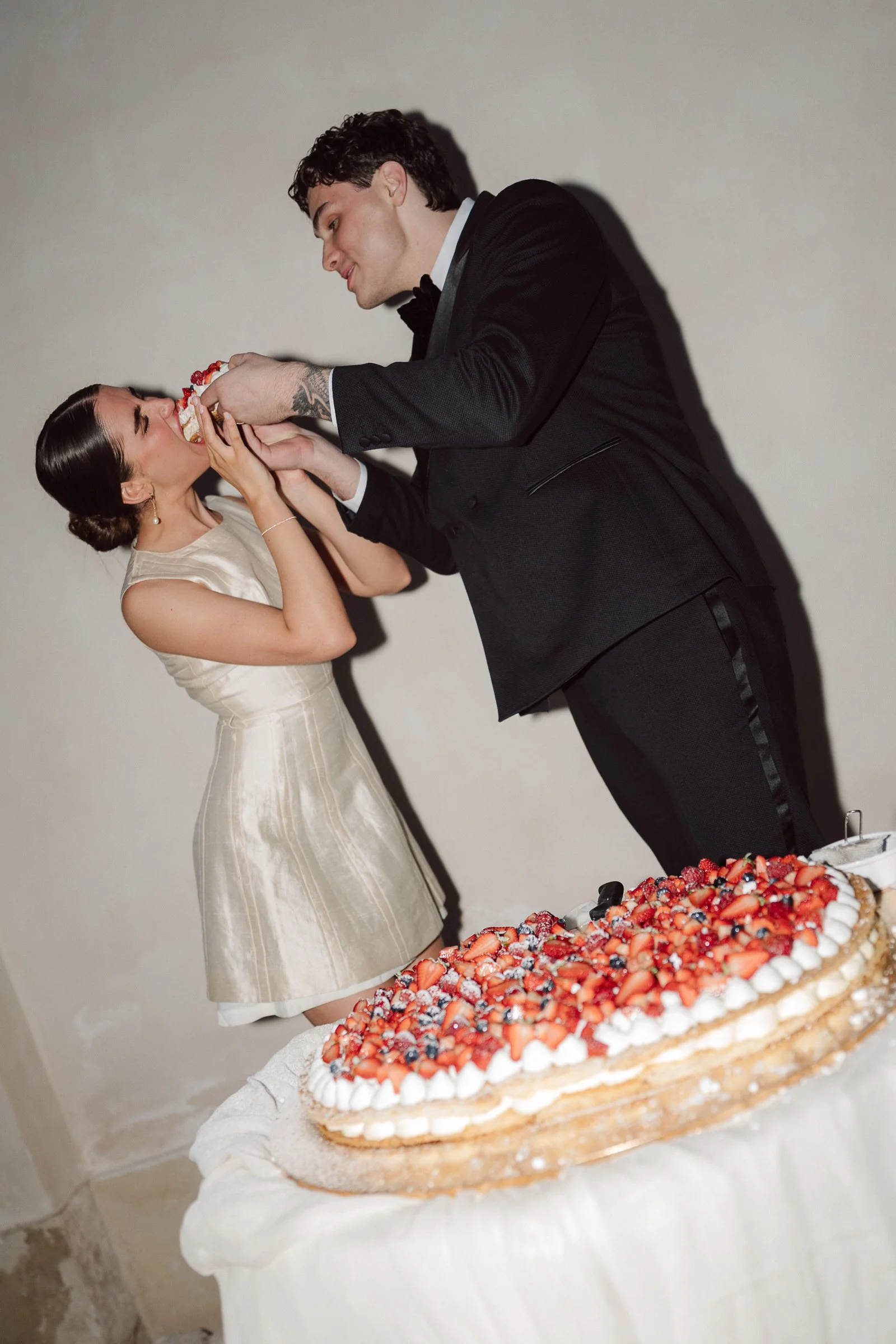 A man in a black tuxedo and a woman in a cream dress share a moment as he feeds her a piece of wedding cake at a celebration, with a large cake decorated with strawberries and other berries in the foreground.