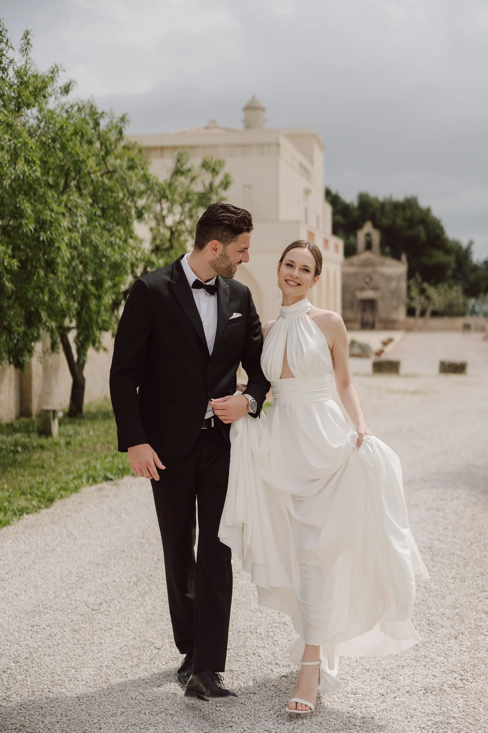 A couple dressed in wedding attire walking outdoors on a gravel path, with ancient buildings and greenery in the background, during a cloudy day.
