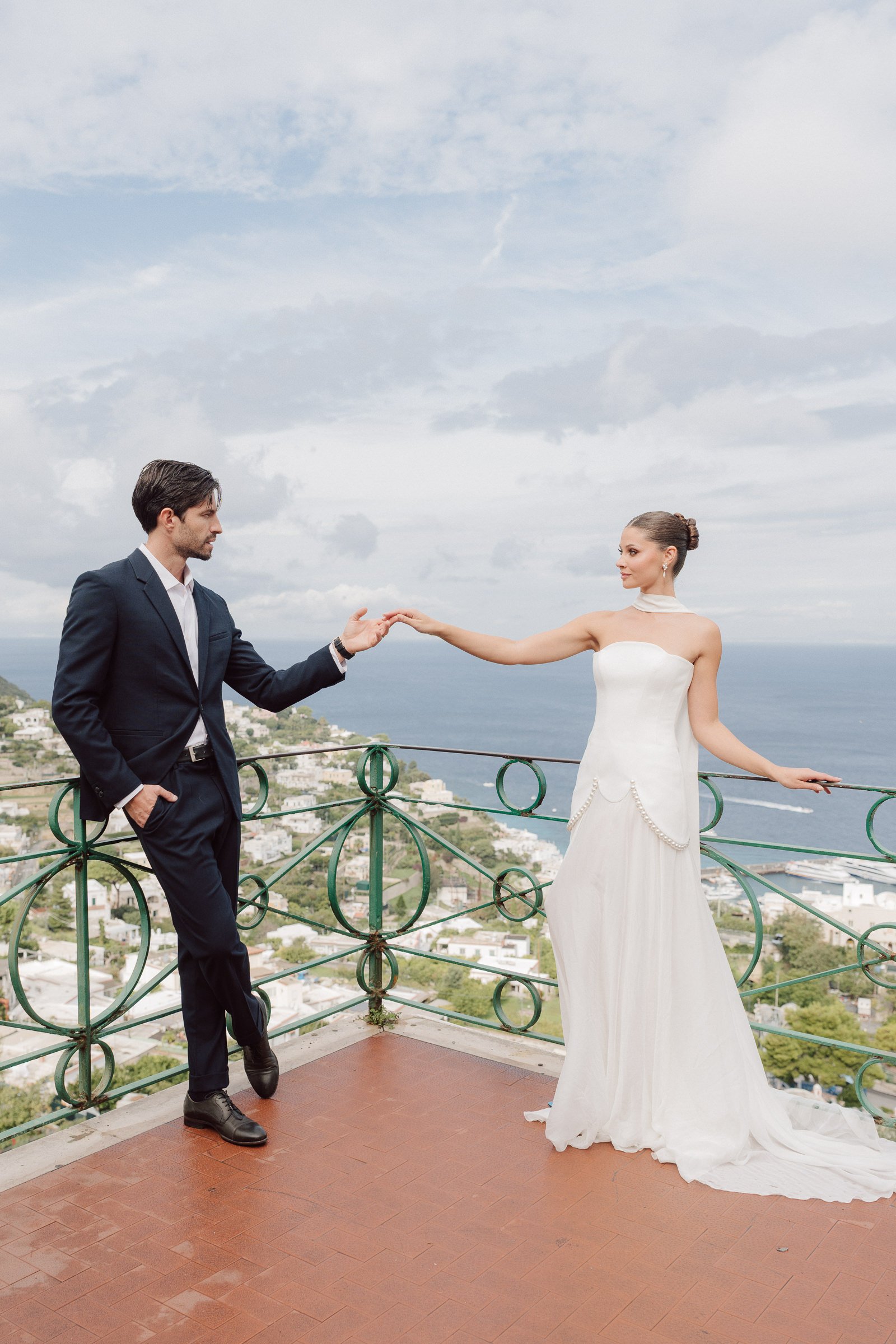 A man in a black suit reaching out his hand to a woman in a white wedding dress on a balcony with ocean and town scenery in the background.