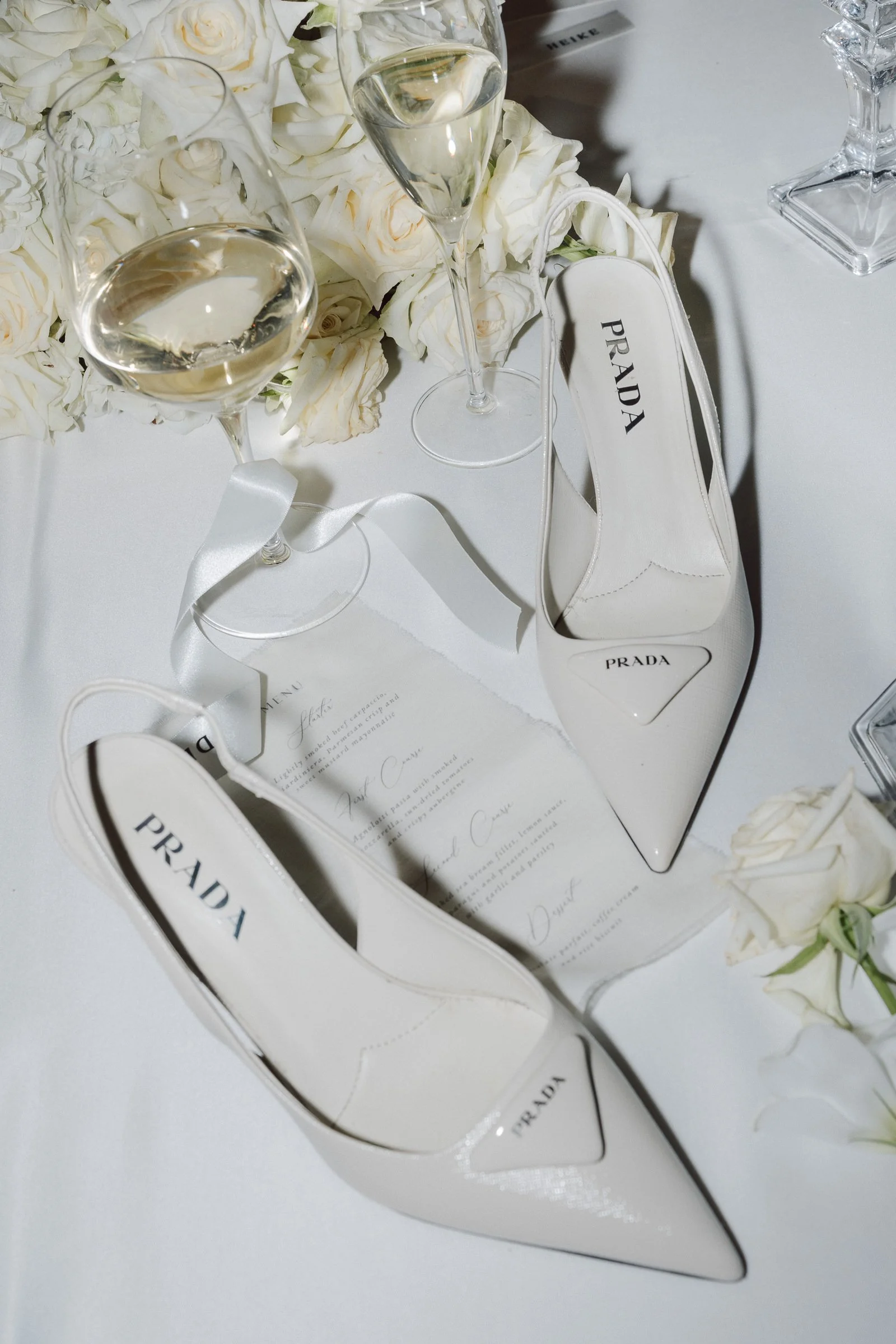 A white Prada high-heeled shoe serving as a jewelry holder on a table with white roses and glasses of champagne.