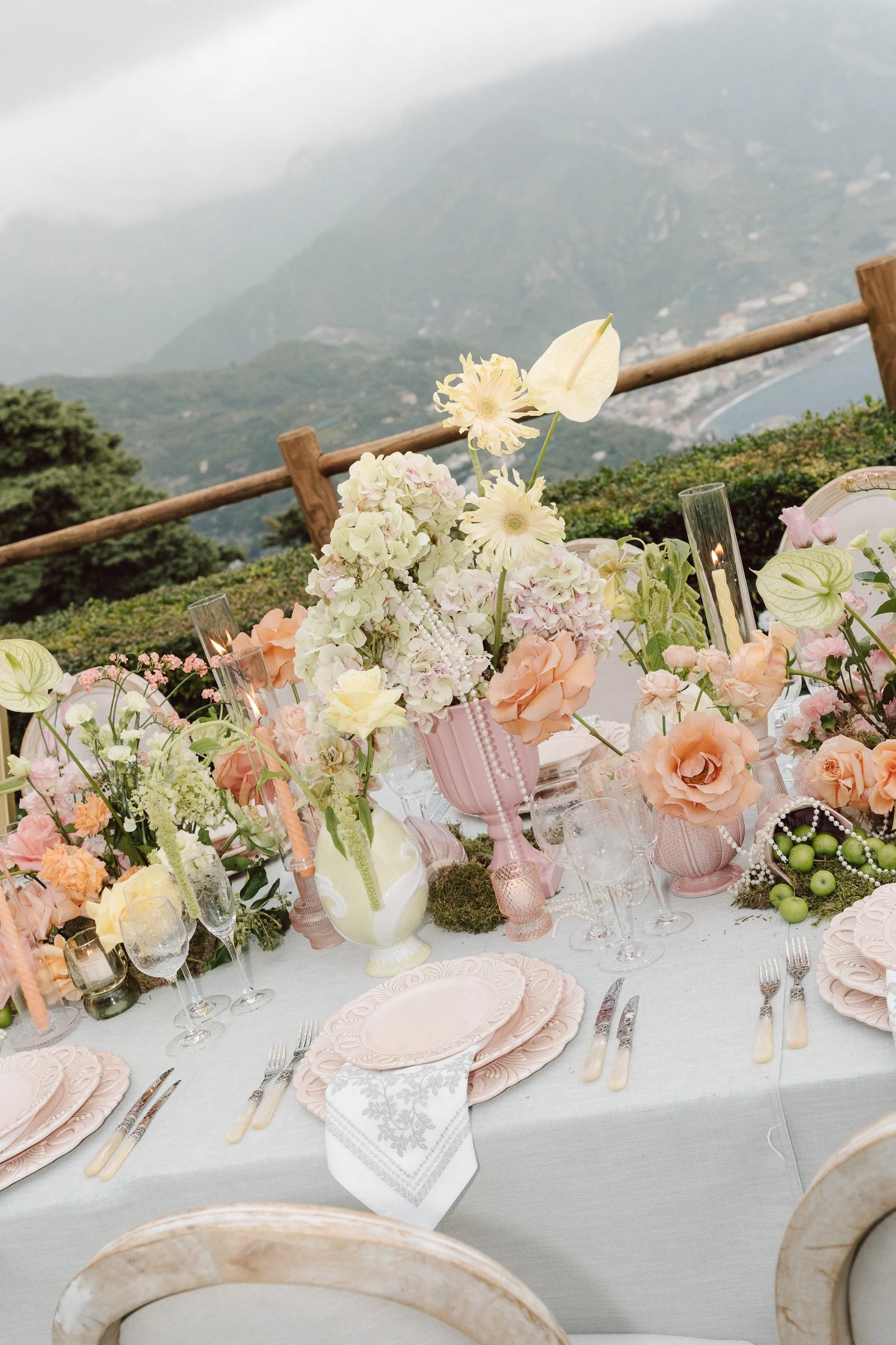 A beautifully decorated outdoor dining table with pastel pink and white floral arrangements, fine china, glassware, and silver cutlery, set against a scenic mountain view with cloudy skies.