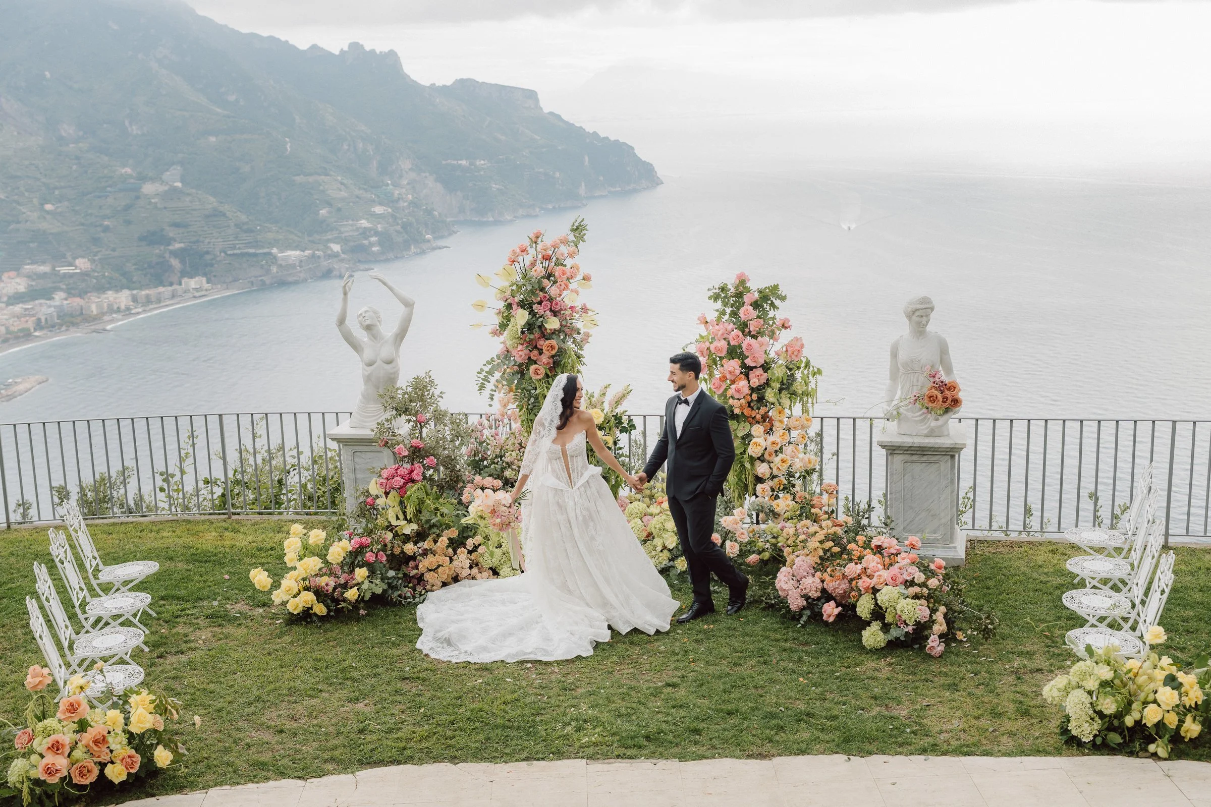 A bride and groom holding hands during a wedding ceremony on a scenic outdoor terrace overlooking a body of water and mountains. The area is decorated with large floral arrangements and classical statues.