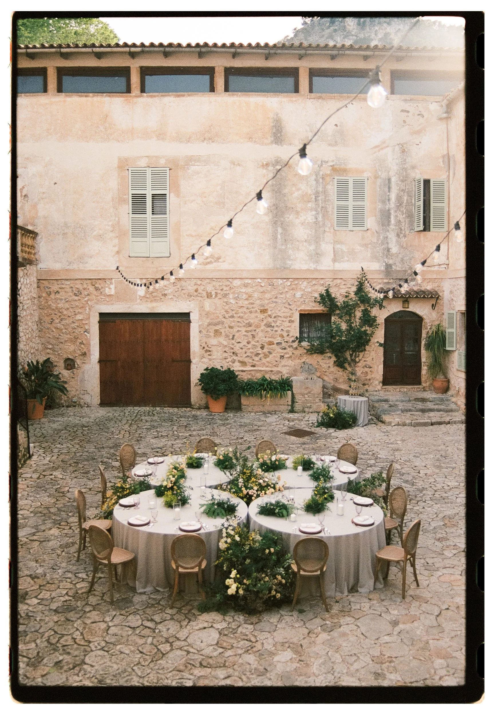 Outdoor courtyard with a round banquet table decorated with floral arrangements, surrounded by chairs, set for a formal event, with stone paving and an old building in the background.