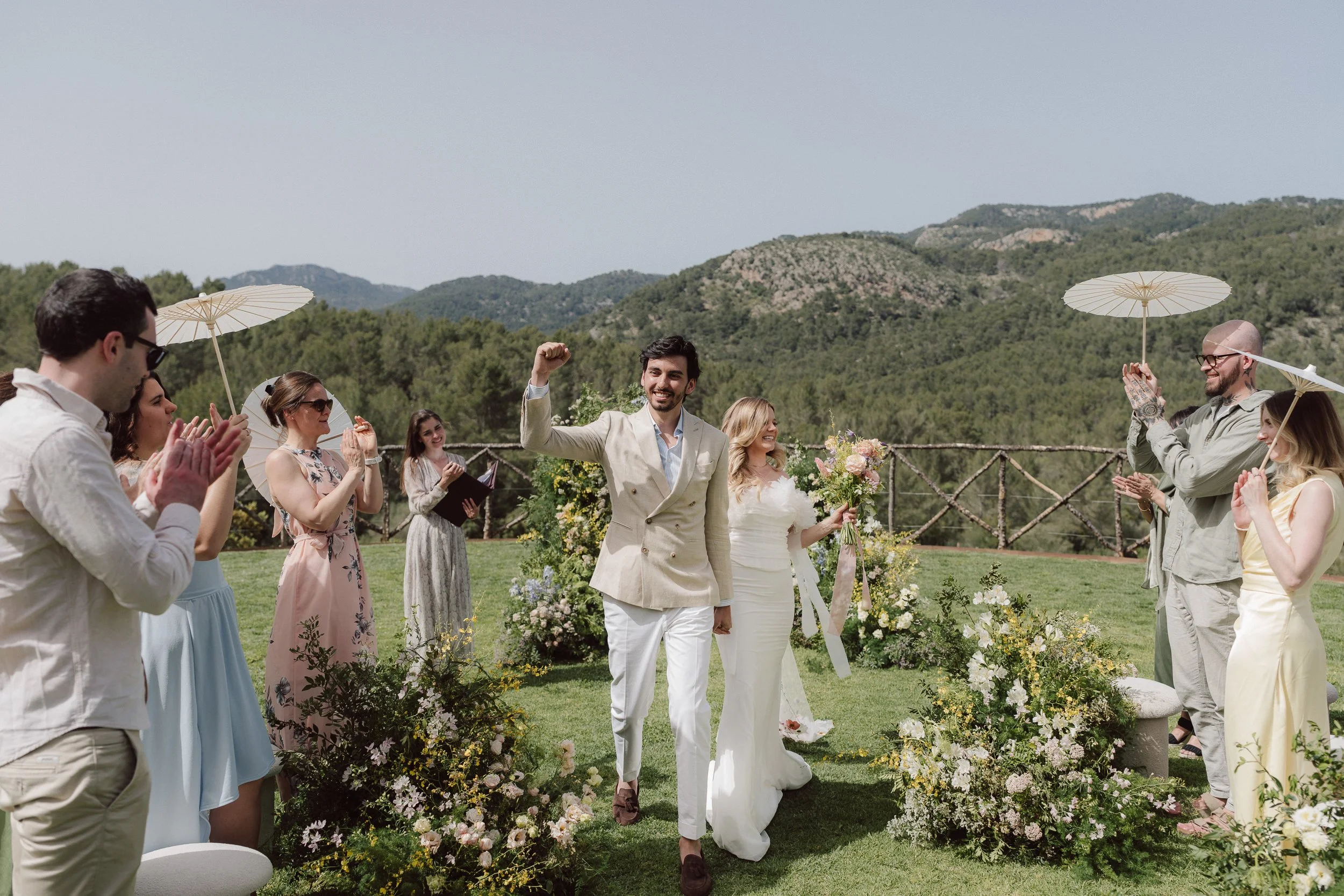 A wedding ceremony outdoors with a groom raising his fist, surrounded by smiling guests holding umbrellas and clapping, set against a backdrop of mountains and greenery.