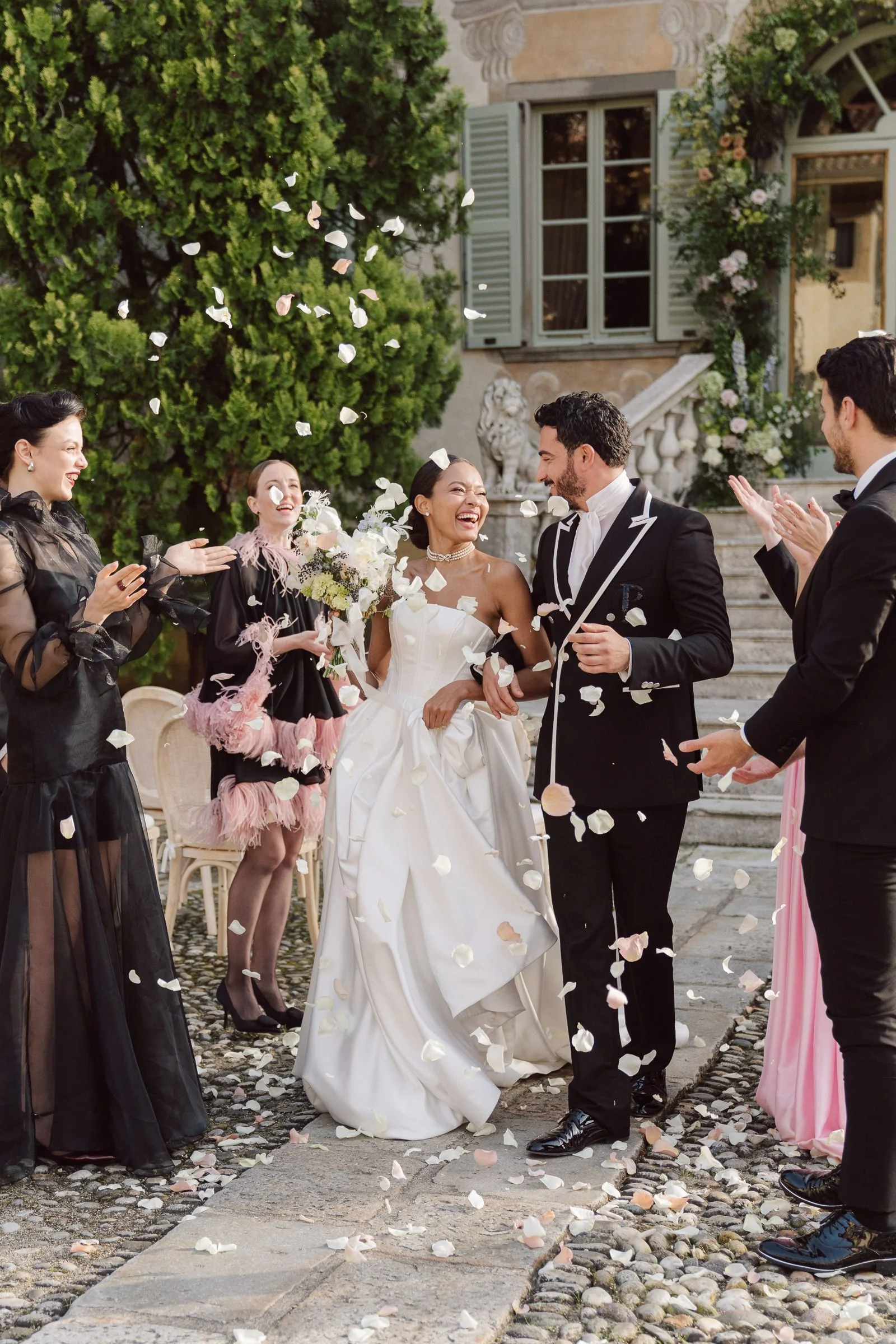 A bride and groom celebrating their wedding outdoors, surrounded by friends. Guests are throwing flower petals and clapping, with a historic building and green trees in the background.