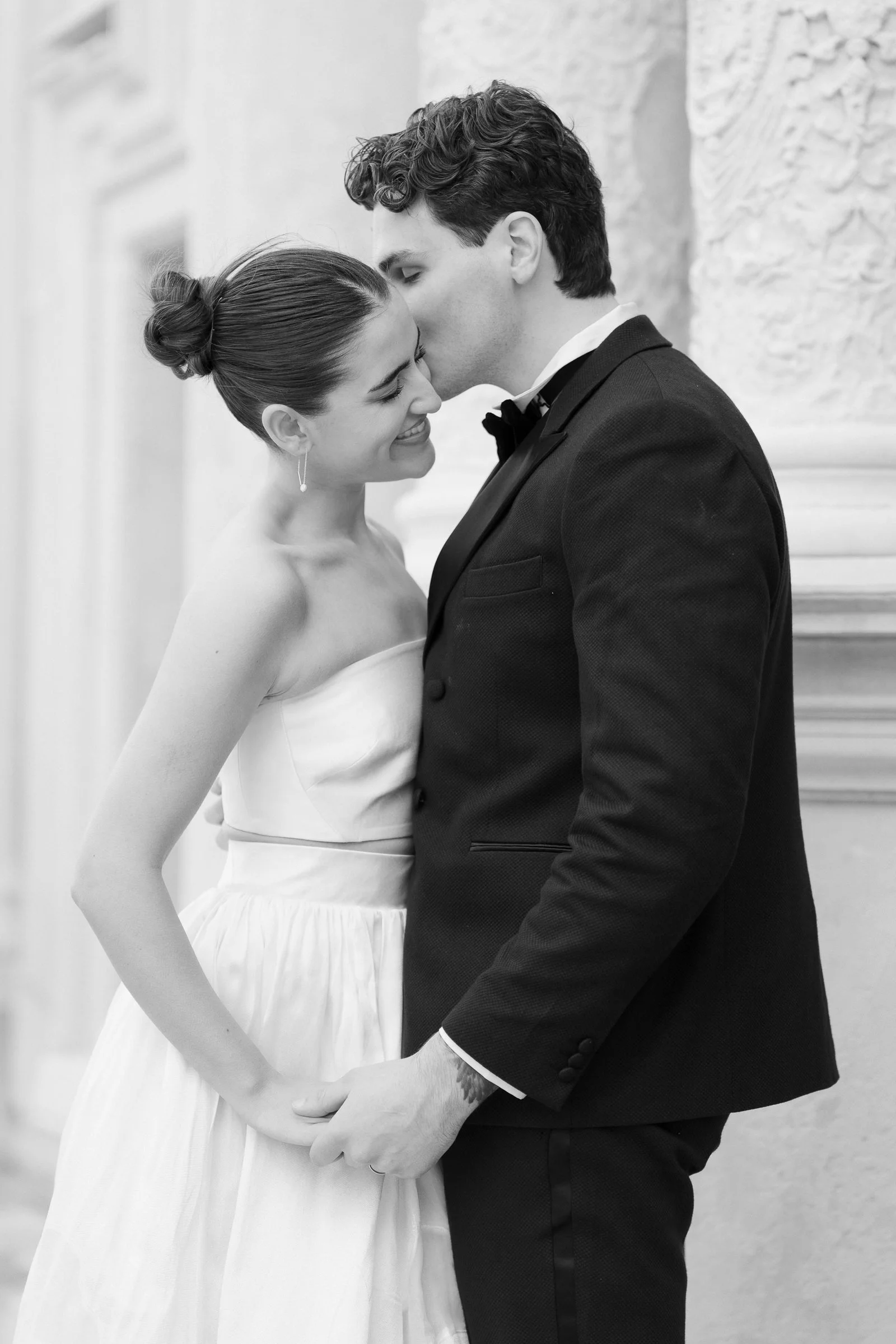 A black-and-white photo of a couple on their wedding day. The bride has her hair in an elegant bun, wearing a strapless wedding dress and earrings. The groom has dark, curly hair, wearing a suit with a bow tie. They are holding hands and touching for