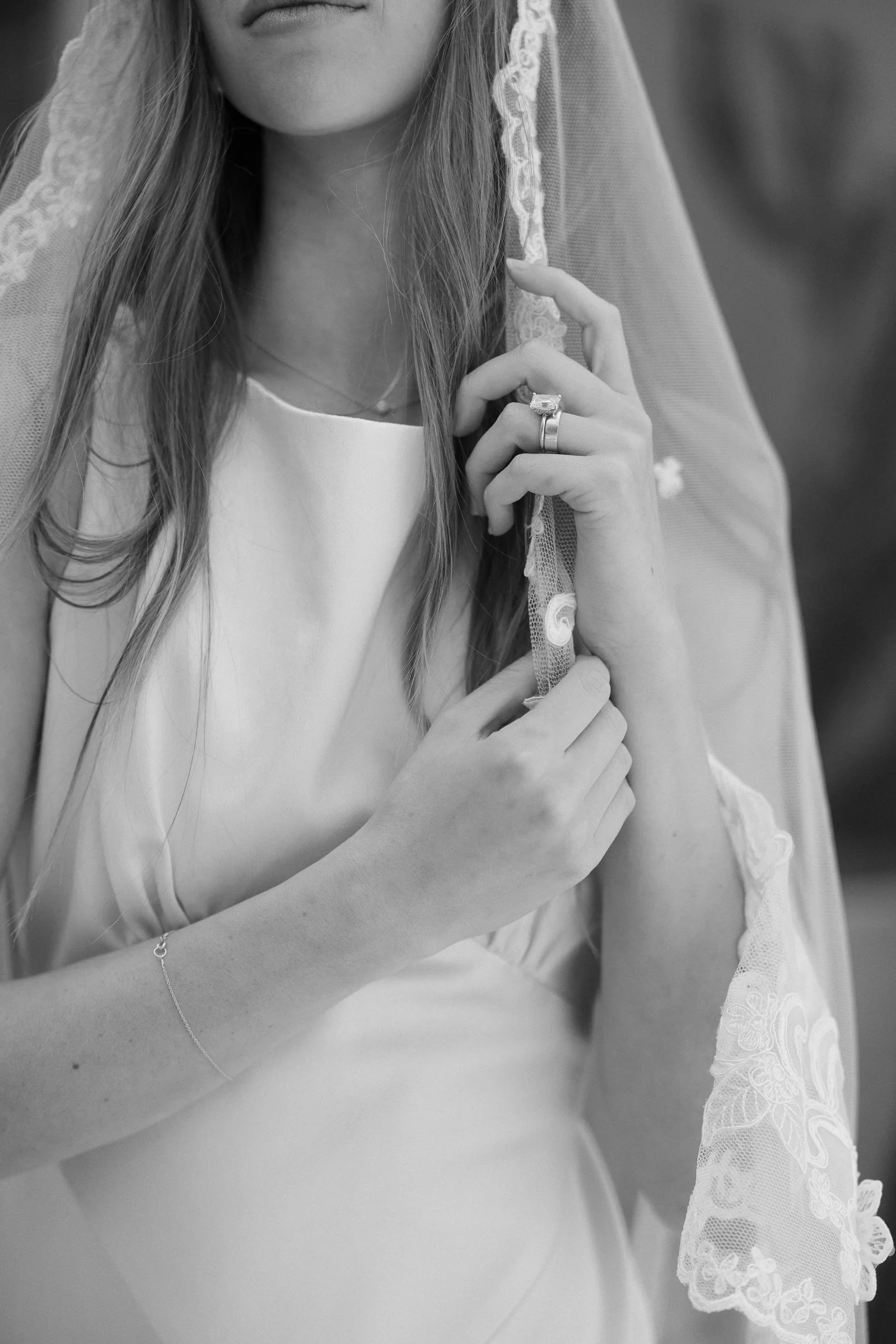 Close-up of a woman in a wedding dress with a lace veil, showing her hands with a wedding ring and jewelry, in black and white.
