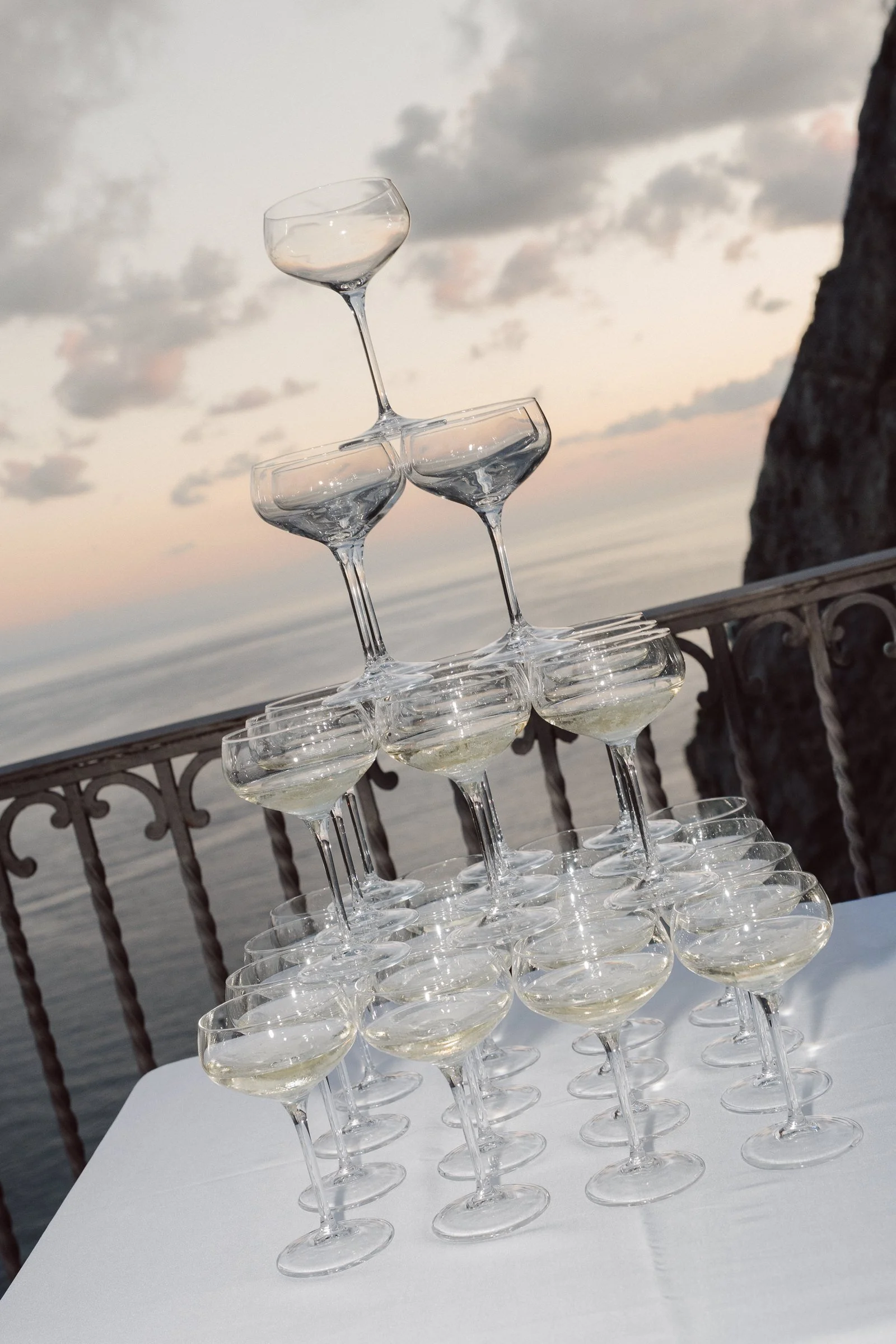Stacked coupe glasses filled with a clear beverage on a white table outdoors, with a cloudy sky and a body of water in the background.