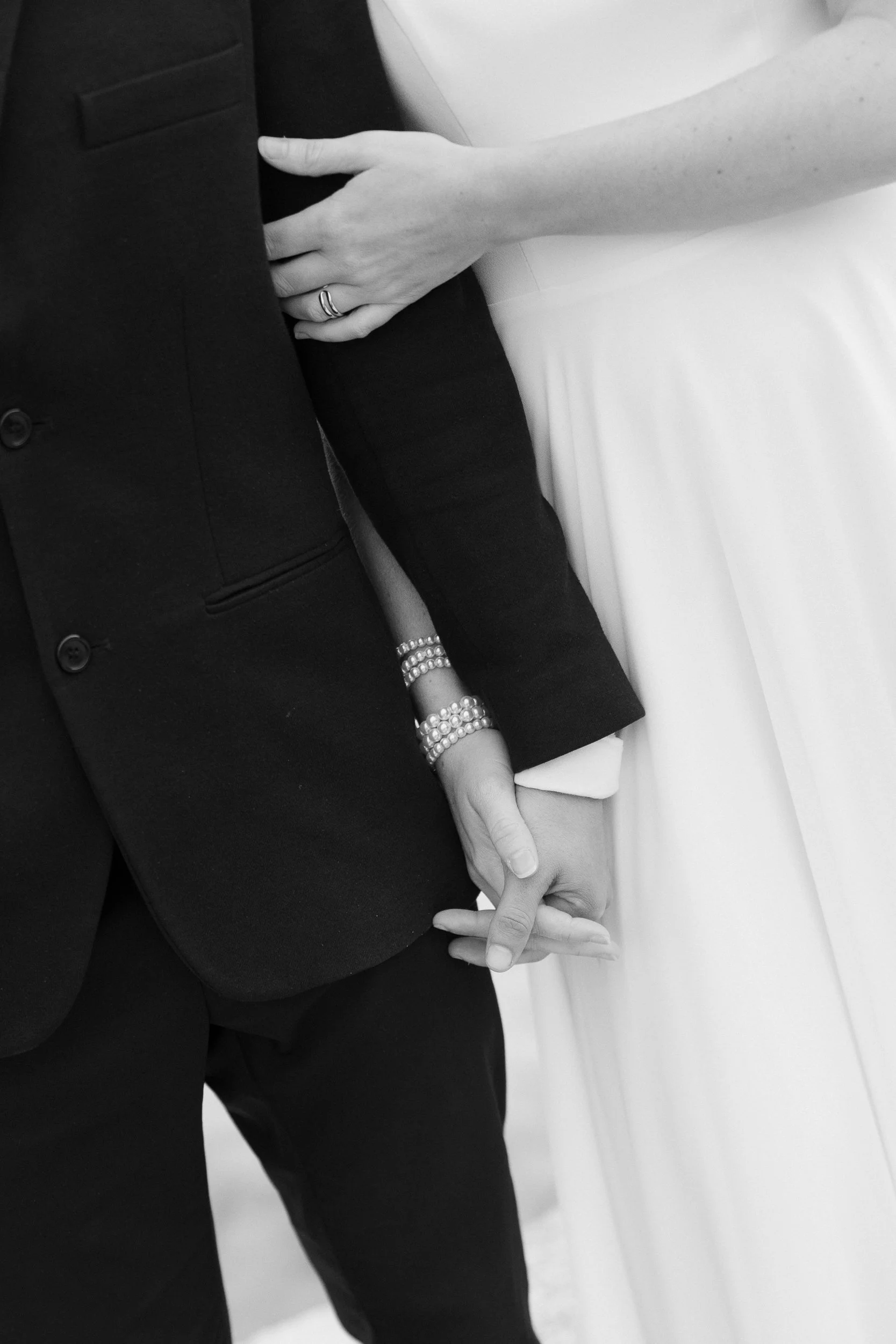 A black and white photo of a couple in formal attire, holding hands with their fingers intertwined, sitting on a bench or couch.