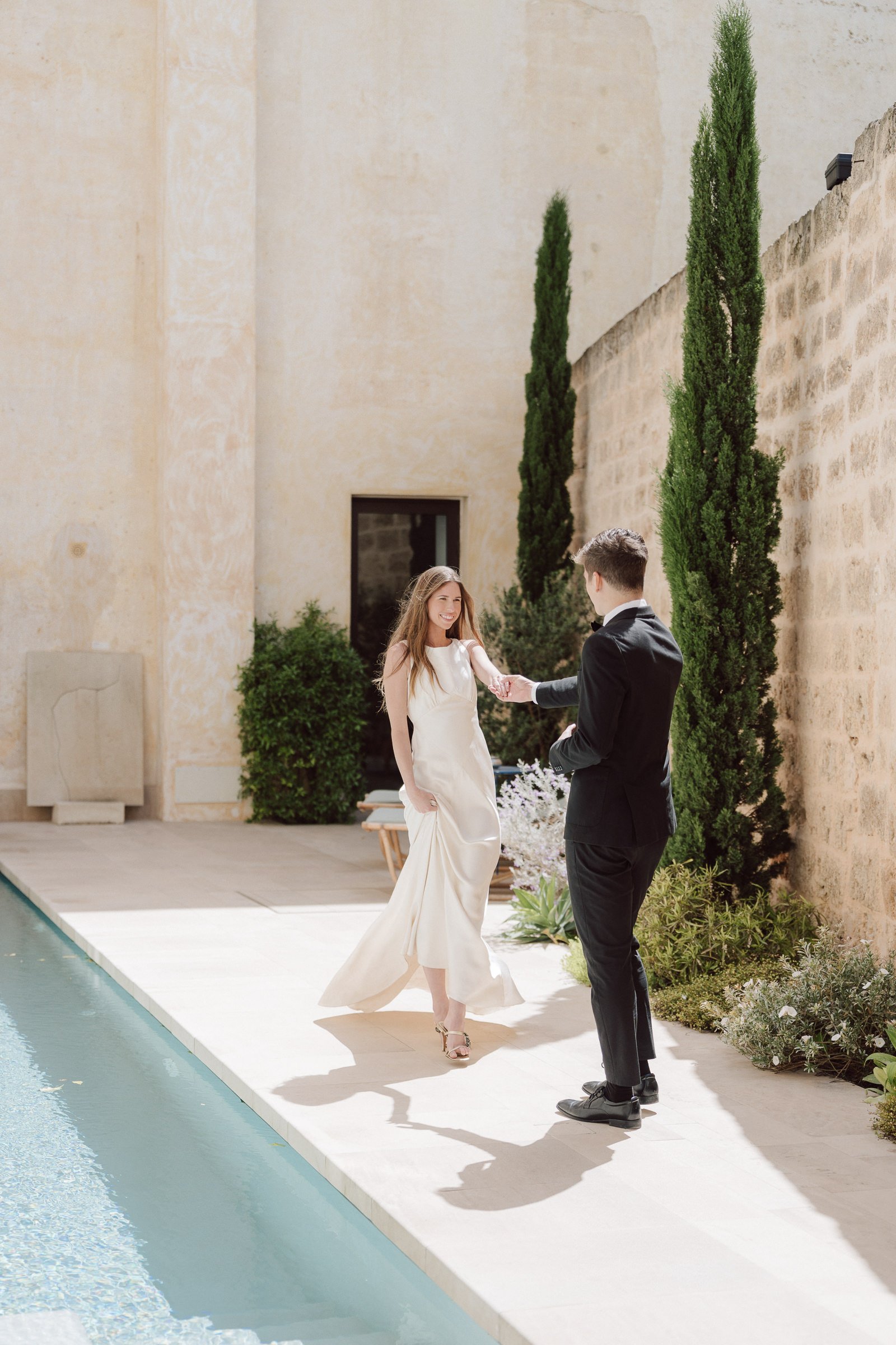 A woman and a man standing by a swimming pool, exchanging rings in an outdoor setting with tall green trees and an old beige stone wall.