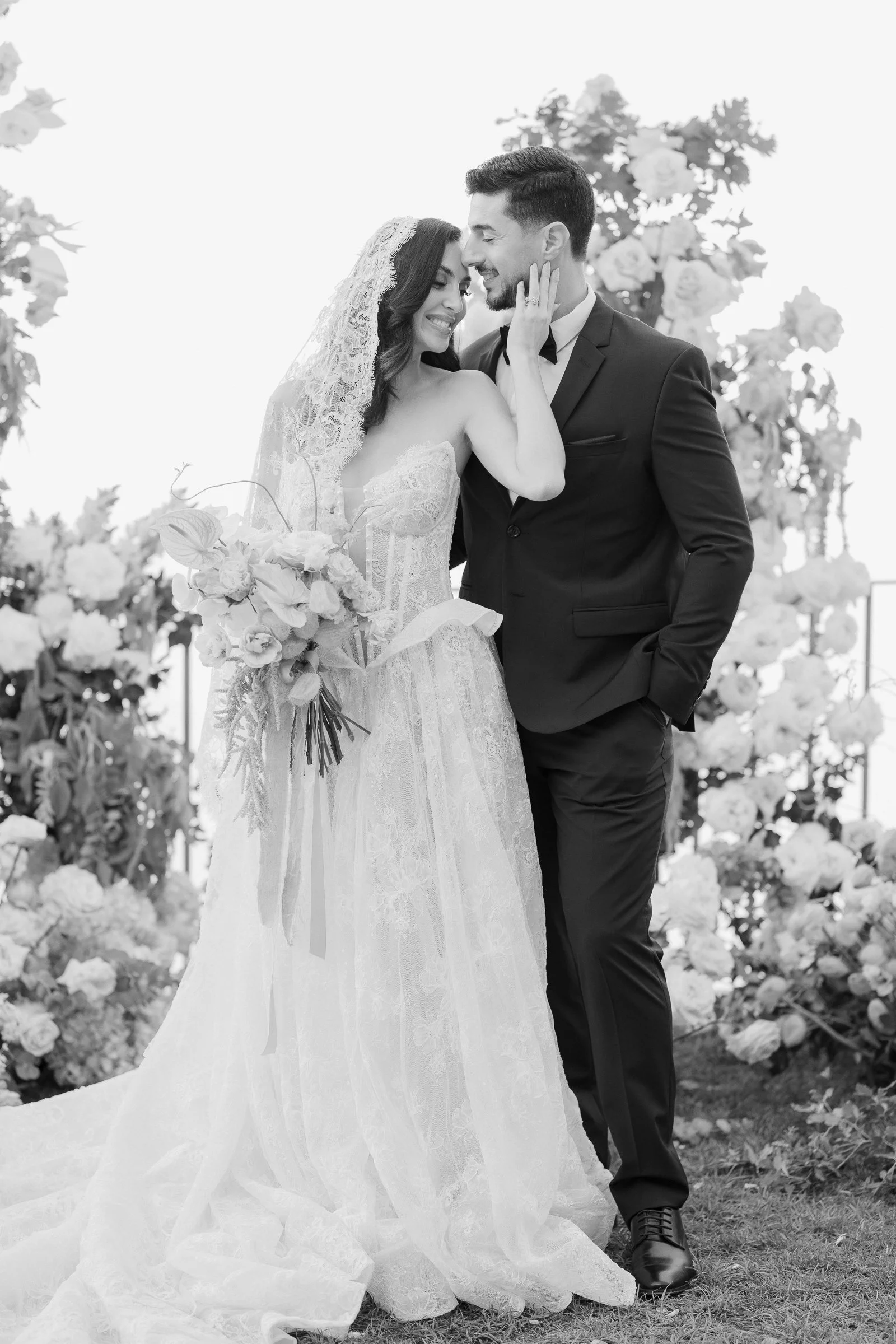 A newlywed couple dressed in formal wedding attire shares a loving moment outdoors, surrounded by flowers and greenery.