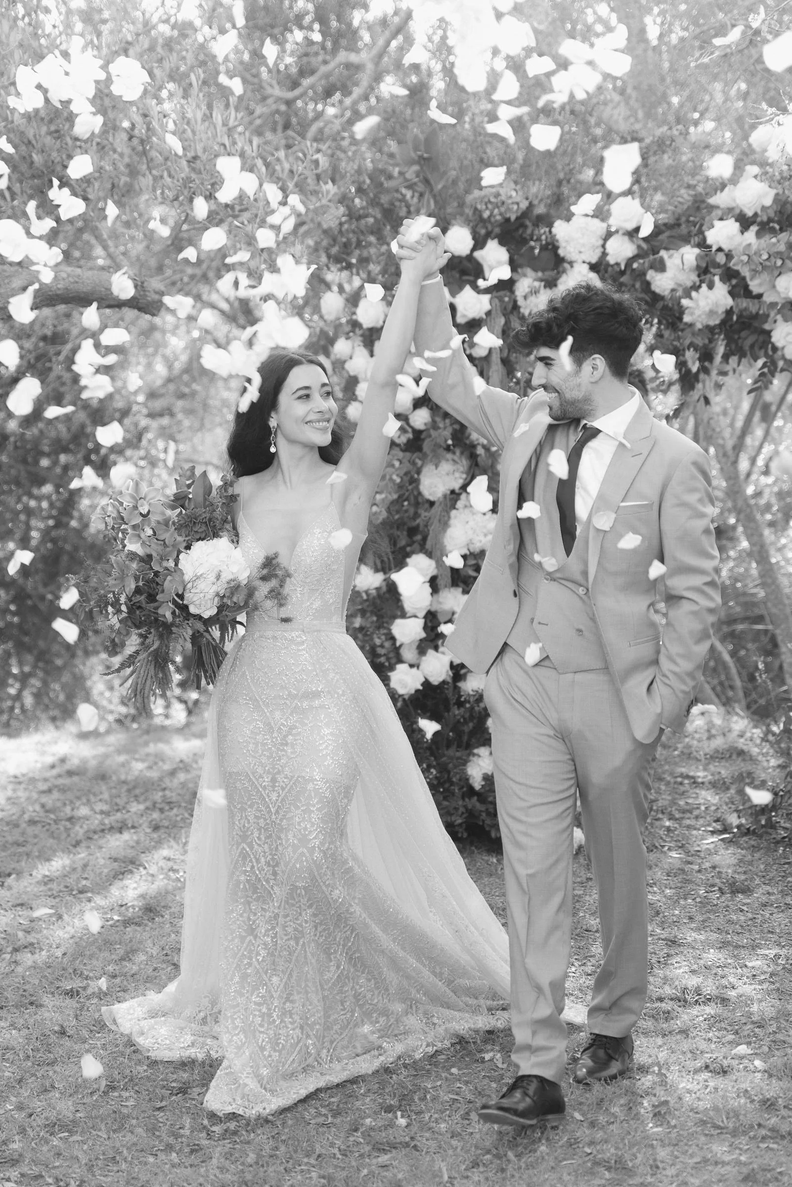 A happy bride and groom outside, holding hands with their arms raised in celebration among falling flower petals, trees, and flowers in the background.