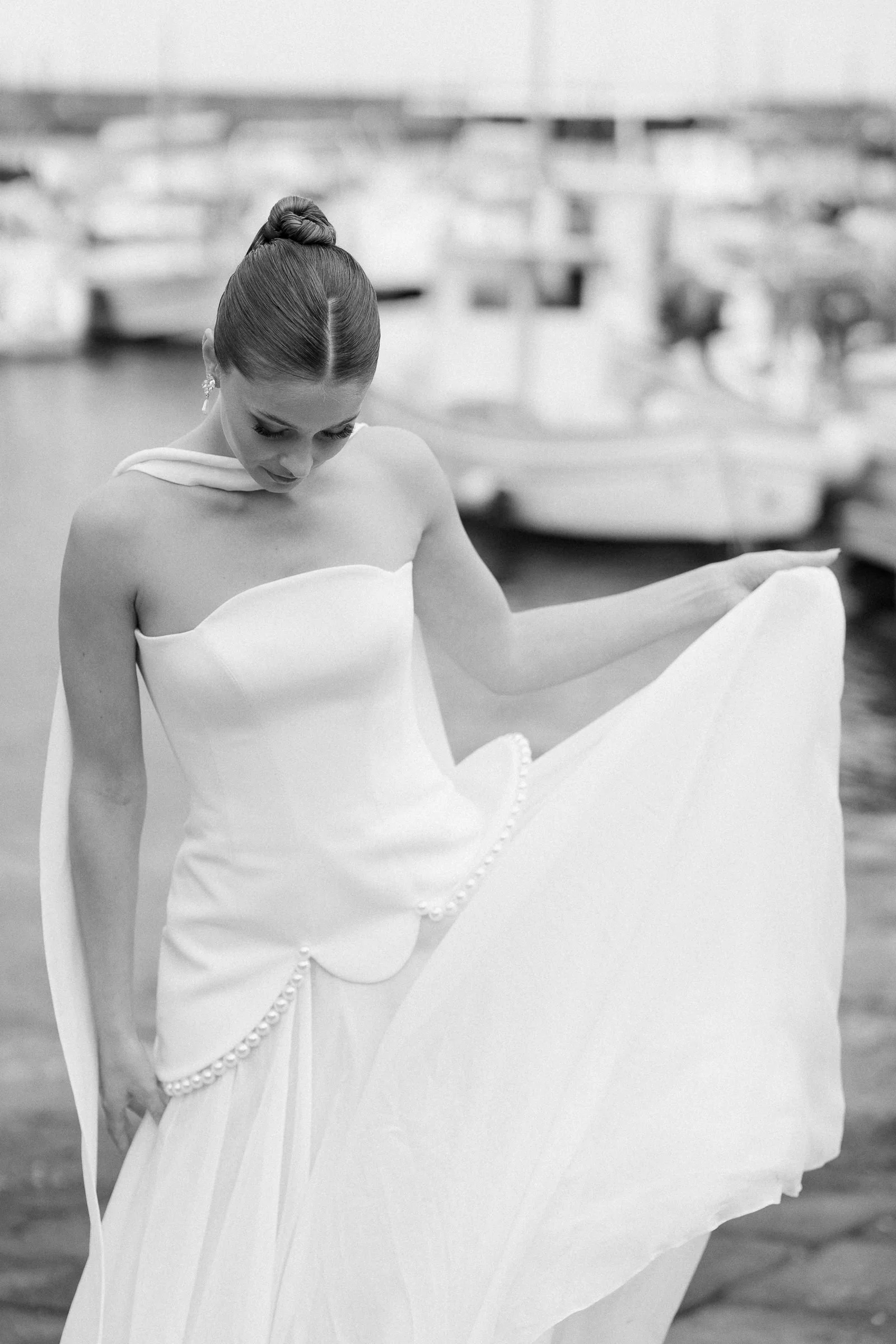 A woman in a white dress with an off-shoulder neckline, holding part of her dress, standing outdoors near a marina with boats and water in the background.